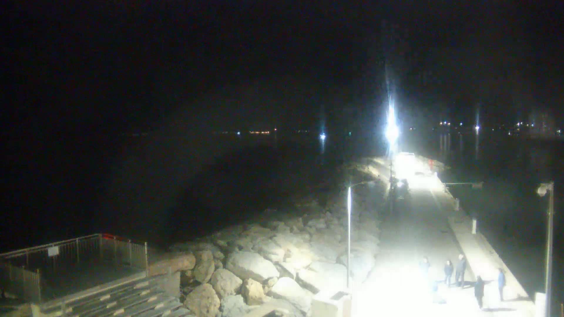 A nighttime view of a paved walkway along a rocky shoreline, with a dark body of water and distant lights visible in the background.