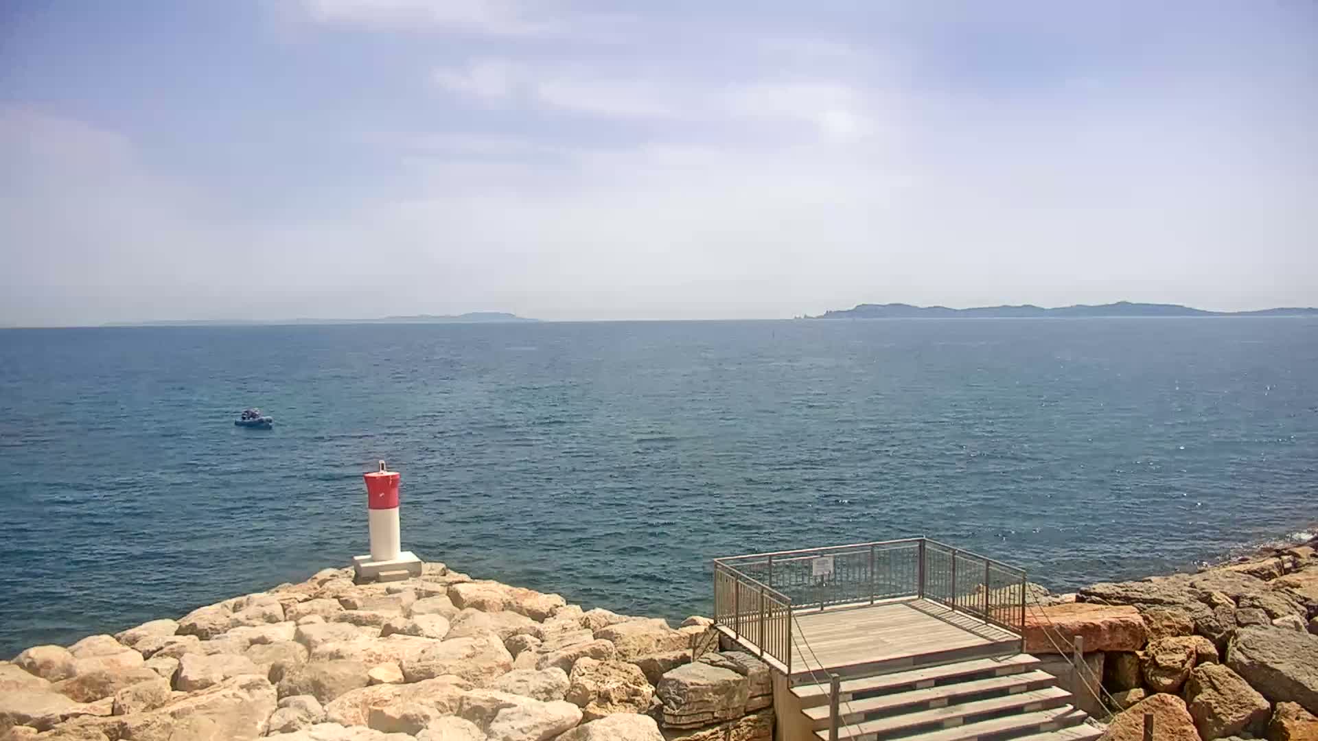 A small boat is visible on a calm, blue sea under a mostly sunny sky, with a red and white marker near a rocky shoreline and a wooden viewing platform with steps.