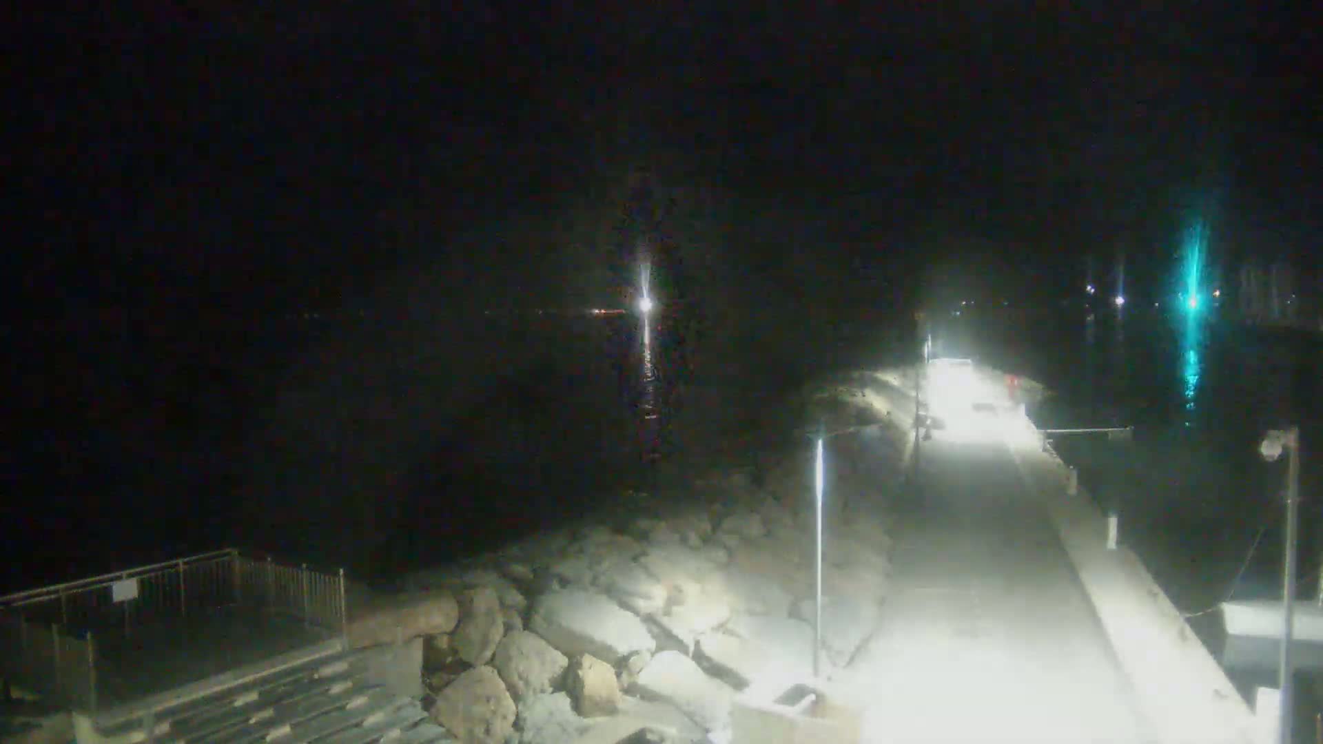 A nighttime view of a paved walkway beside a body of water, with rocky embankments and lights illuminating the scene.