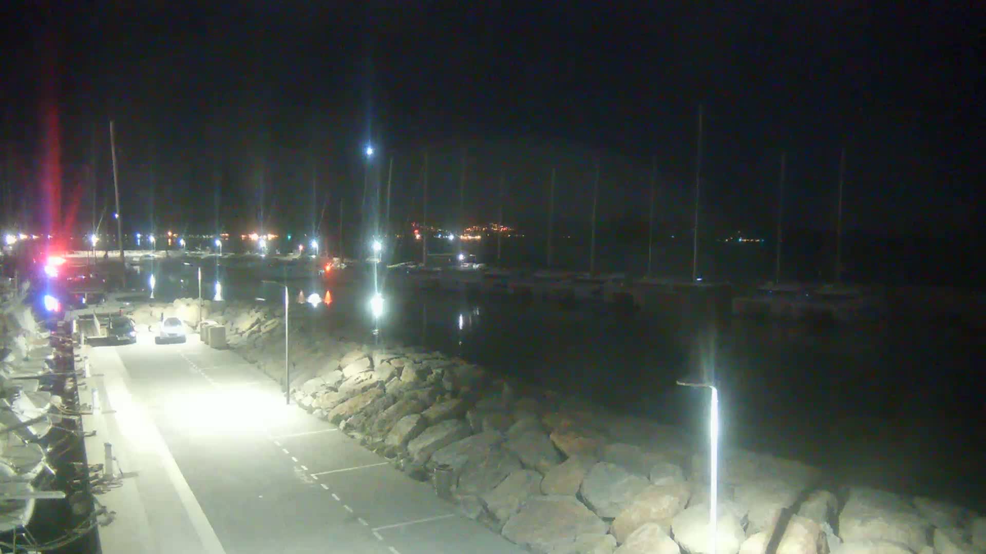 A nighttime view of a harbor with docked boats and a paved roadway lined with large rocks, illuminated by artificial lights.