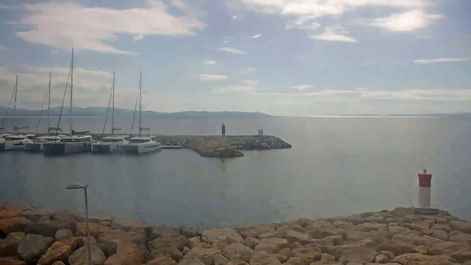 Several sailboats are docked in a calm harbor on a partly cloudy day, with a rocky shoreline and a distant landmass visible.