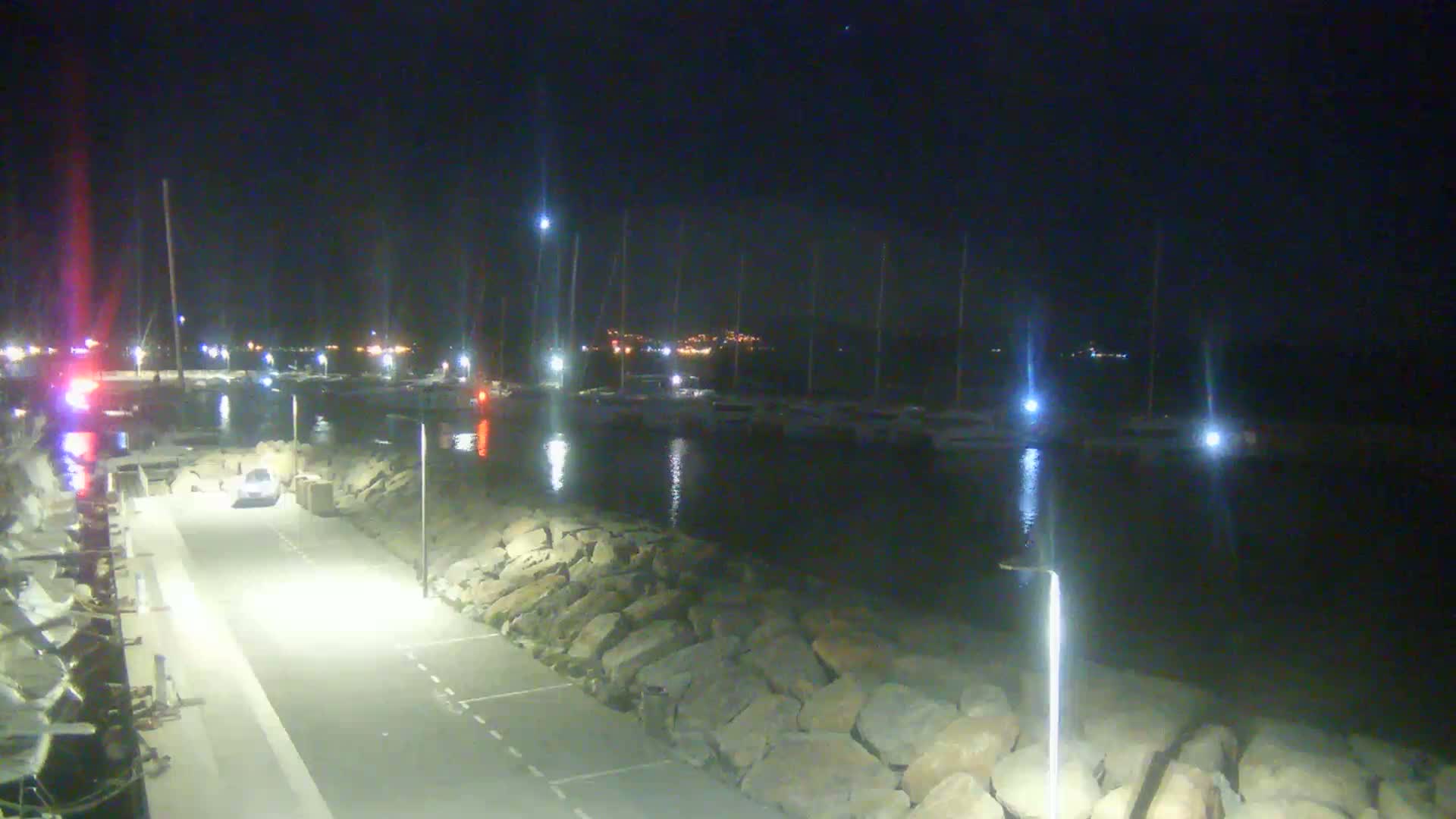 A nighttime view of a harbor with docked sailboats, a roadway with a single car, and a rocky shoreline illuminated by artificial lights.
