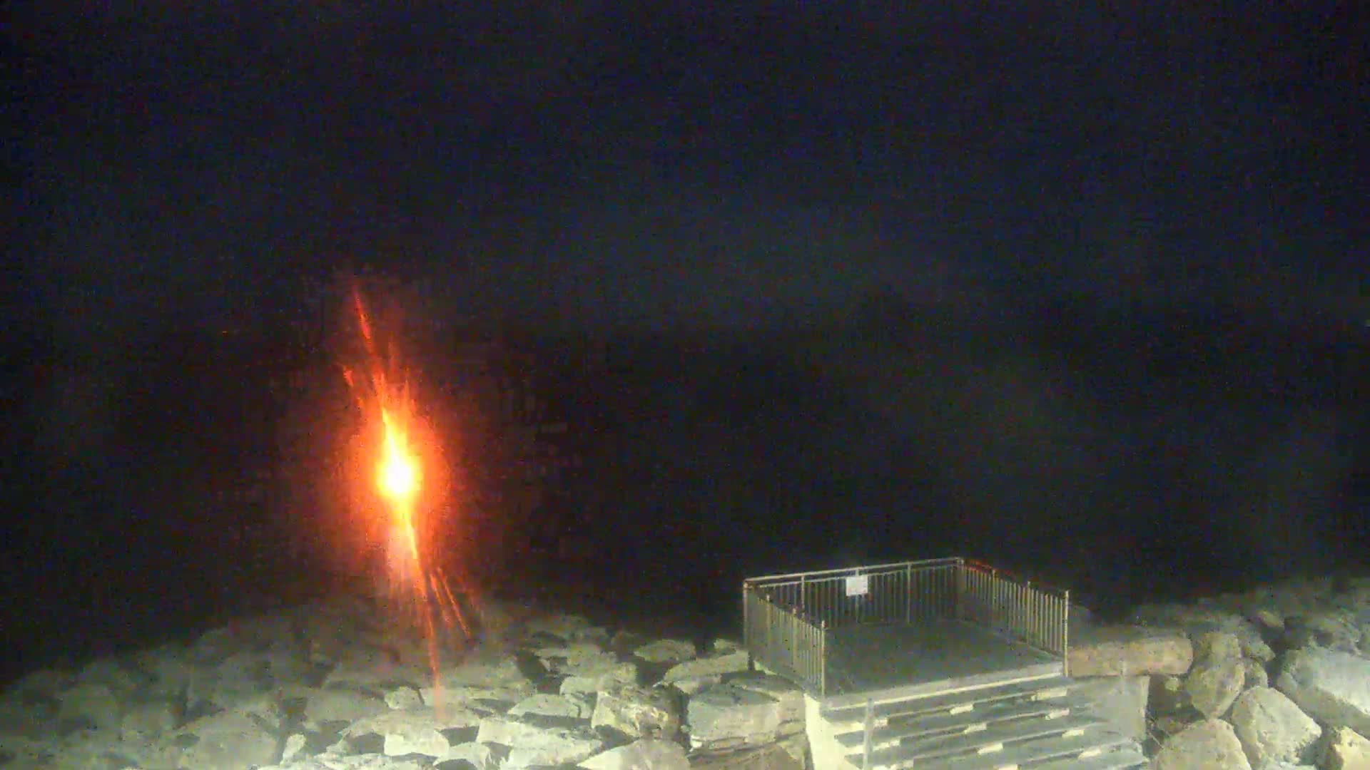 A bright orange light shines over a rocky shoreline at night near a set of stairs and a railing.