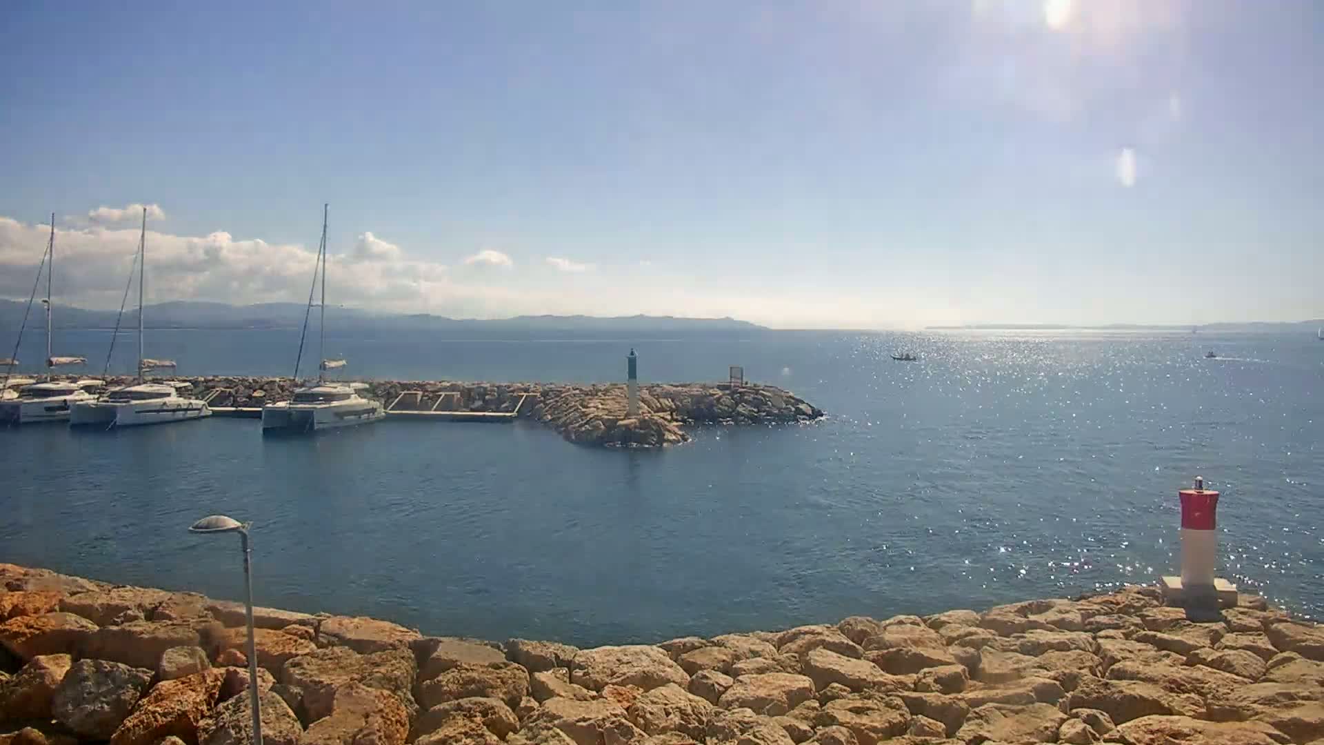 Several sailboats are moored in a calm harbor on a sunny day with a few scattered clouds.
