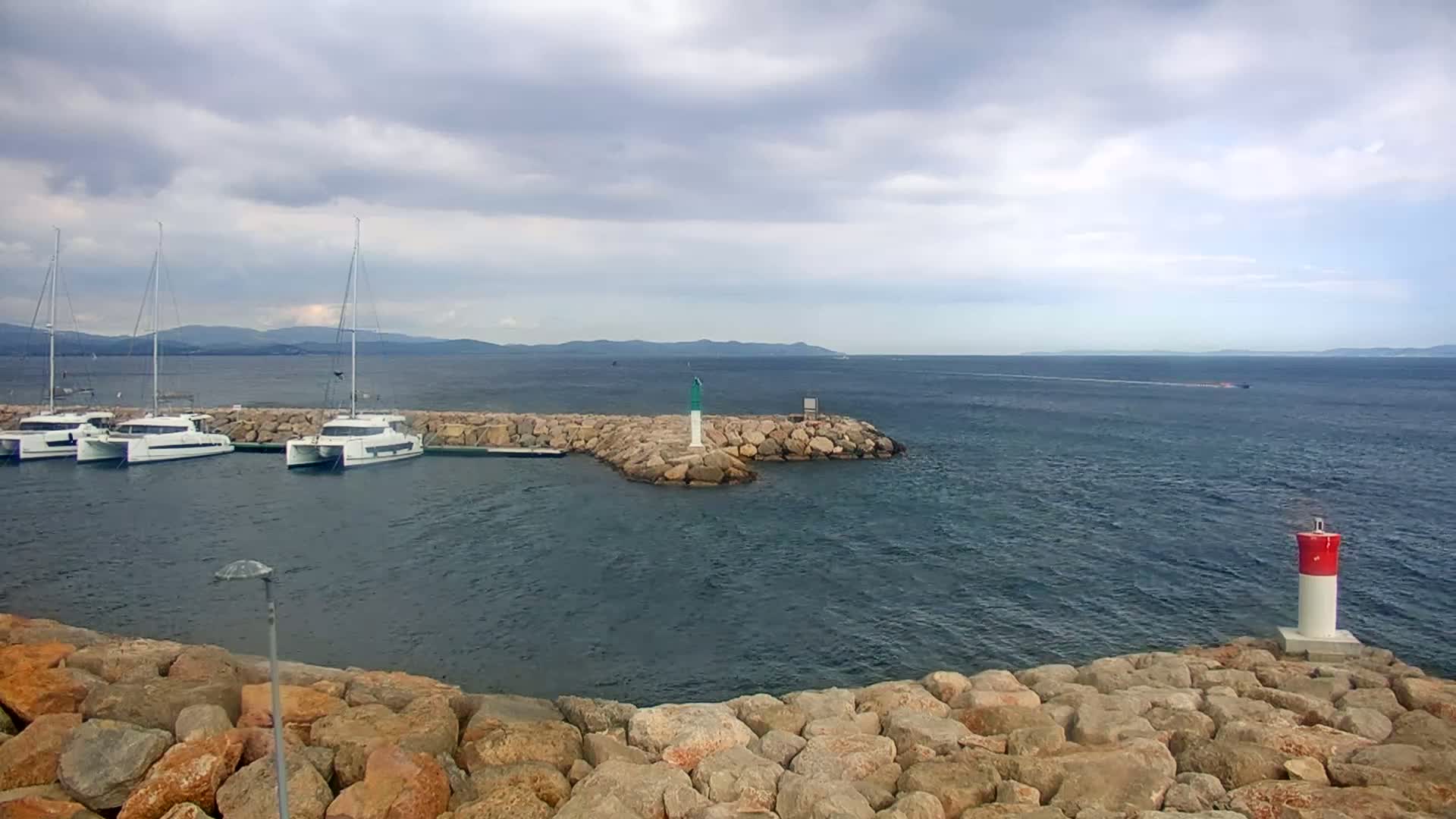 Several white catamarans are moored in a calm harbor protected by a rock breakwater under a cloudy sky, with distant mountains visible across the water.