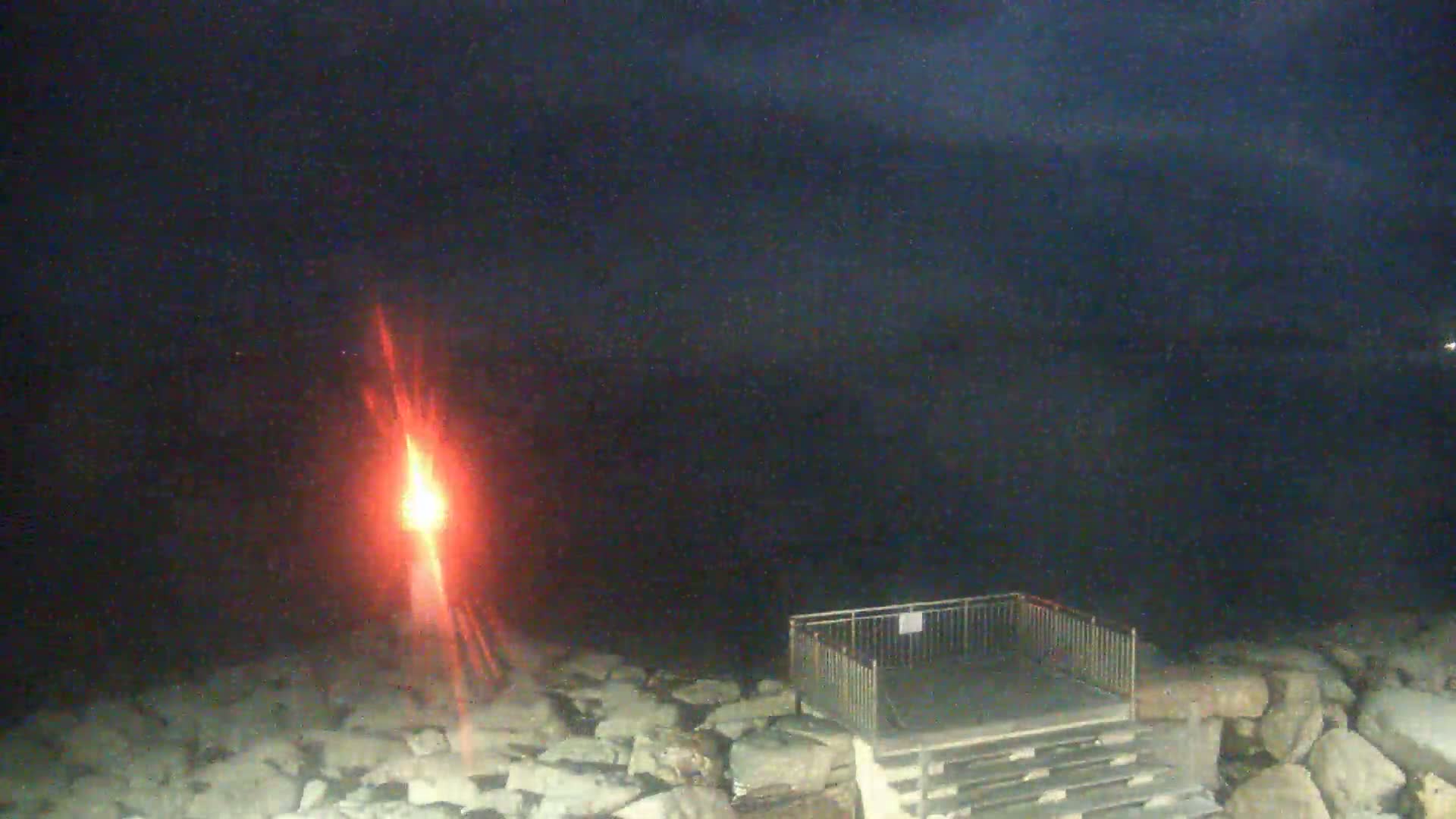 A bright red light shines near a stone shoreline at night, with a small fenced-in platform and steps visible nearby.