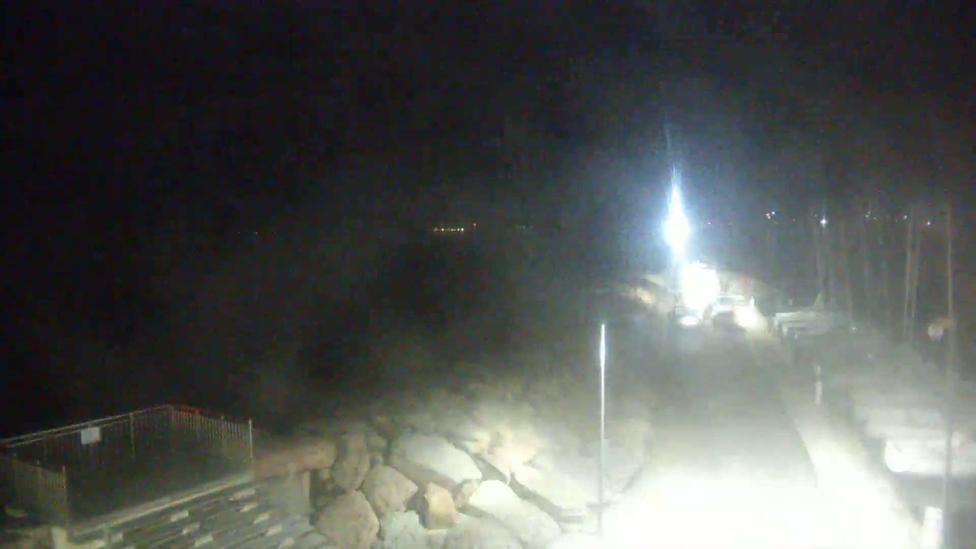 A nighttime view shows a brightly lit paved area near a body of water, with rocky terrain and parked boats visible in the distance.