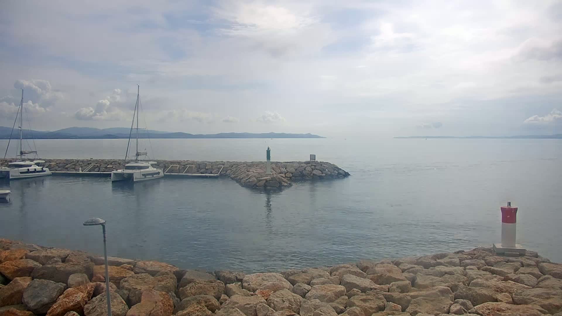 Two sailboats are moored in a calm harbor, with a rocky breakwater separating the harbor from the open sea under a partly cloudy sky.