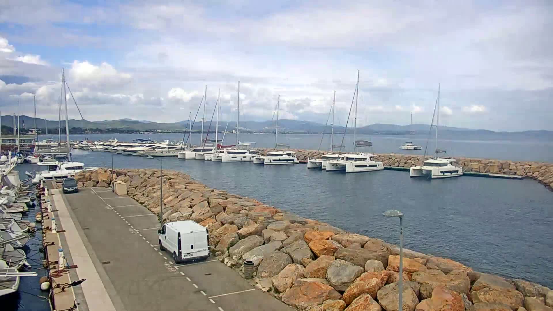 A marina with many sailboats and motor yachts is nestled behind a rock wall on a partly cloudy day.