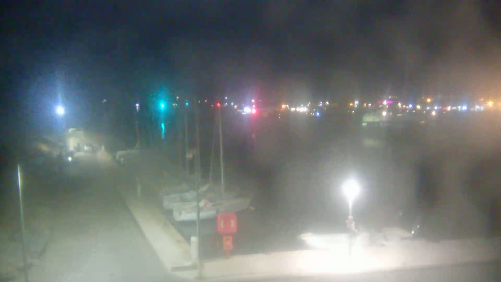 A nighttime view of a harbor with docked sailboats and faint lights from buildings and vehicles in the background, under hazy or foggy conditions.