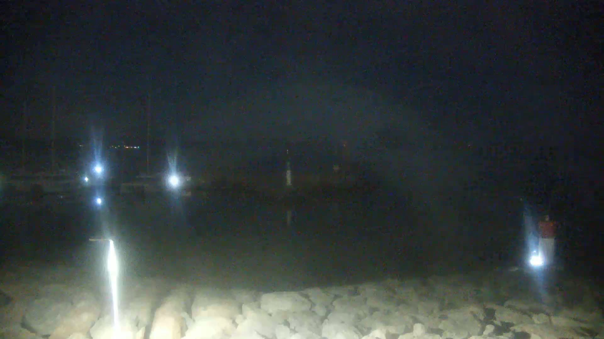 A dark, nighttime scene shows a body of water with several lights reflecting on the surface, boats in the distance, and a rocky shoreline.
