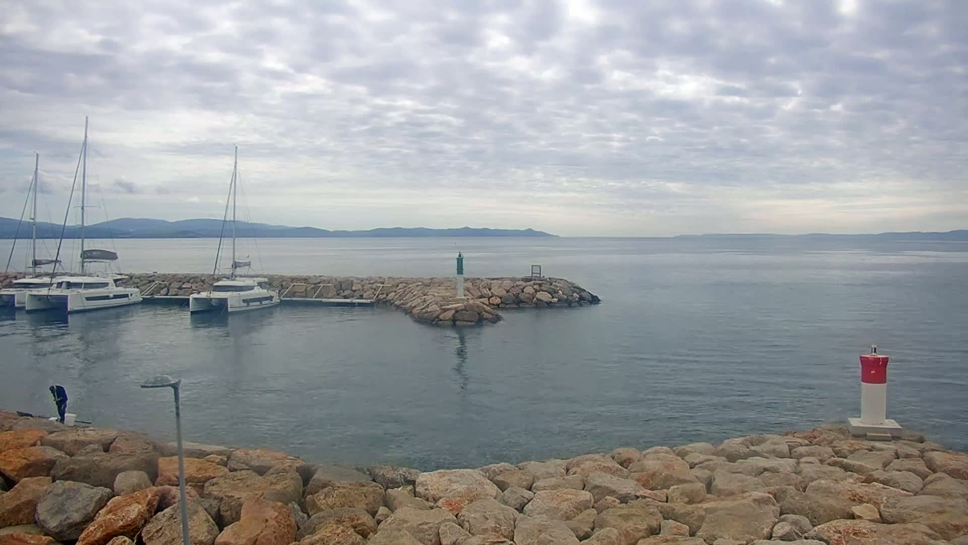 Three sailboats are moored in a calm harbor on a cloudy day, with a rocky breakwater and distant mountains visible.