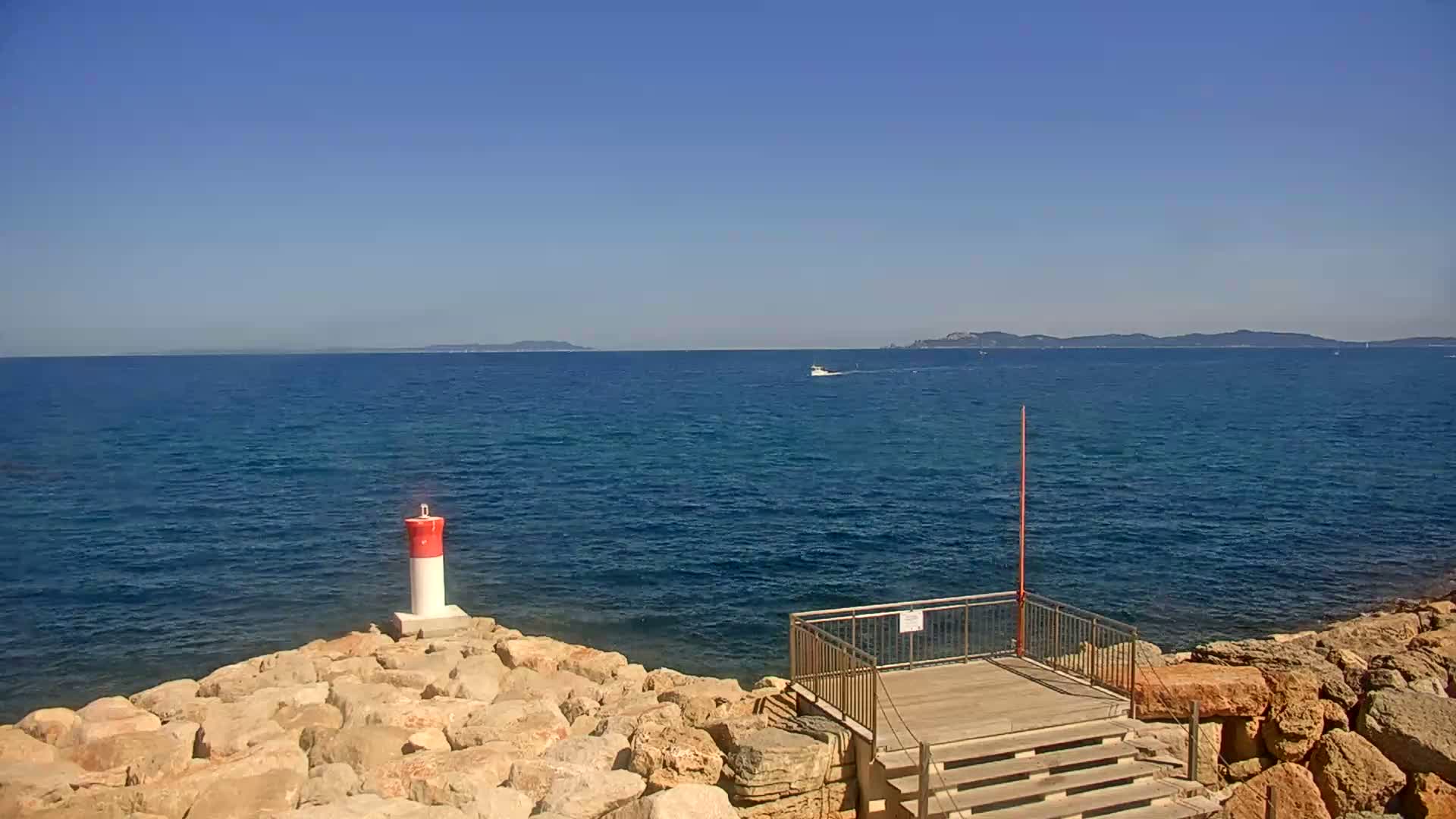 A small, red-and-white lighthouse stands on a rocky shore overlooking a calm blue sea under a clear blue sky.