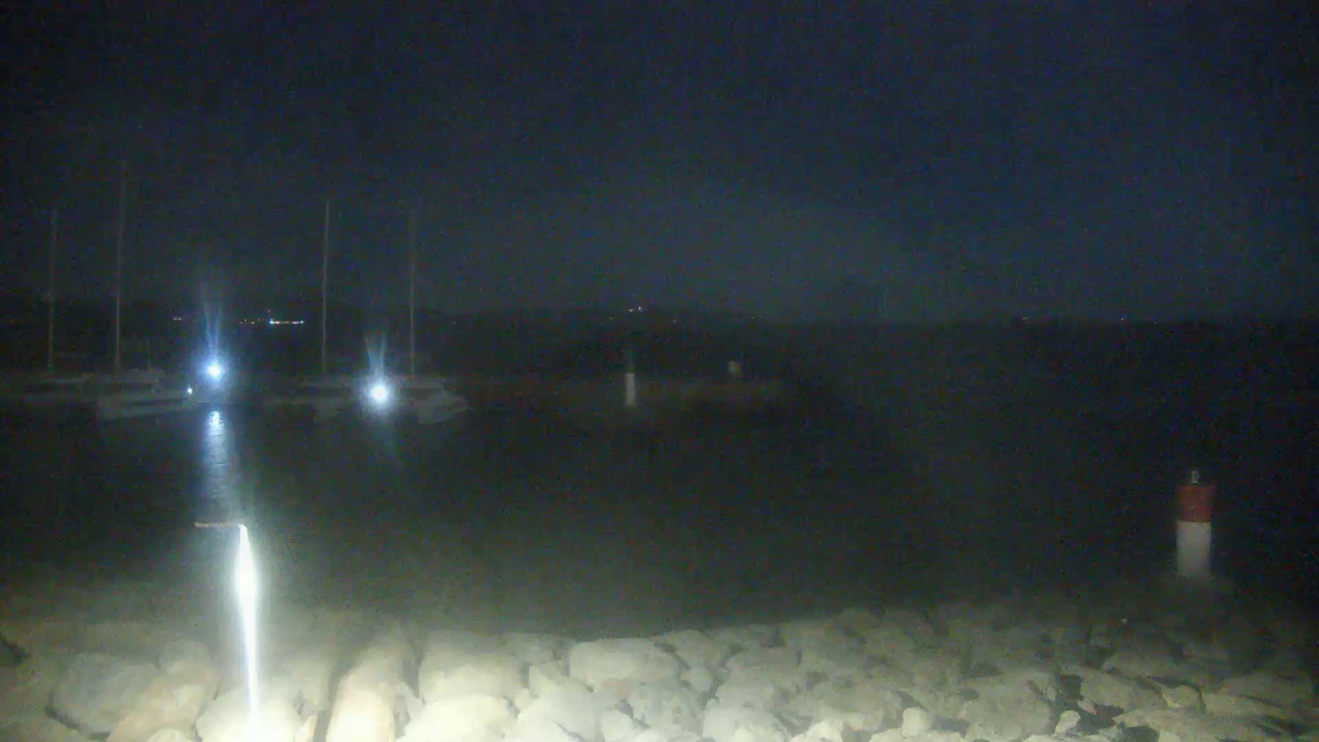 At night, a dark body of water is visible with several boats docked, illuminated by lights, and a rocky shoreline in the foreground.