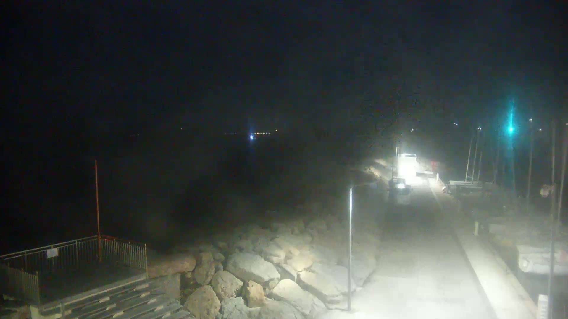 A nighttime view shows a road next to a rocky shoreline with a body of water in the background, lit by streetlights.