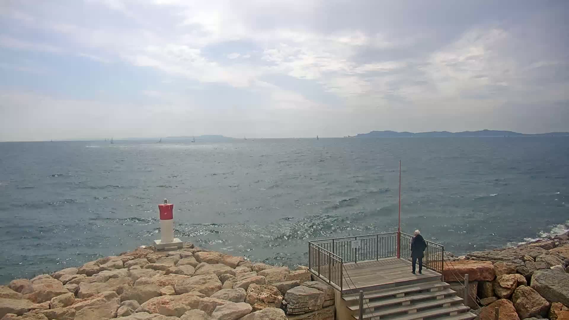 A person stands on a wooden platform overlooking a rocky shoreline and a somewhat choppy sea with distant sailboats under a partly cloudy sky.