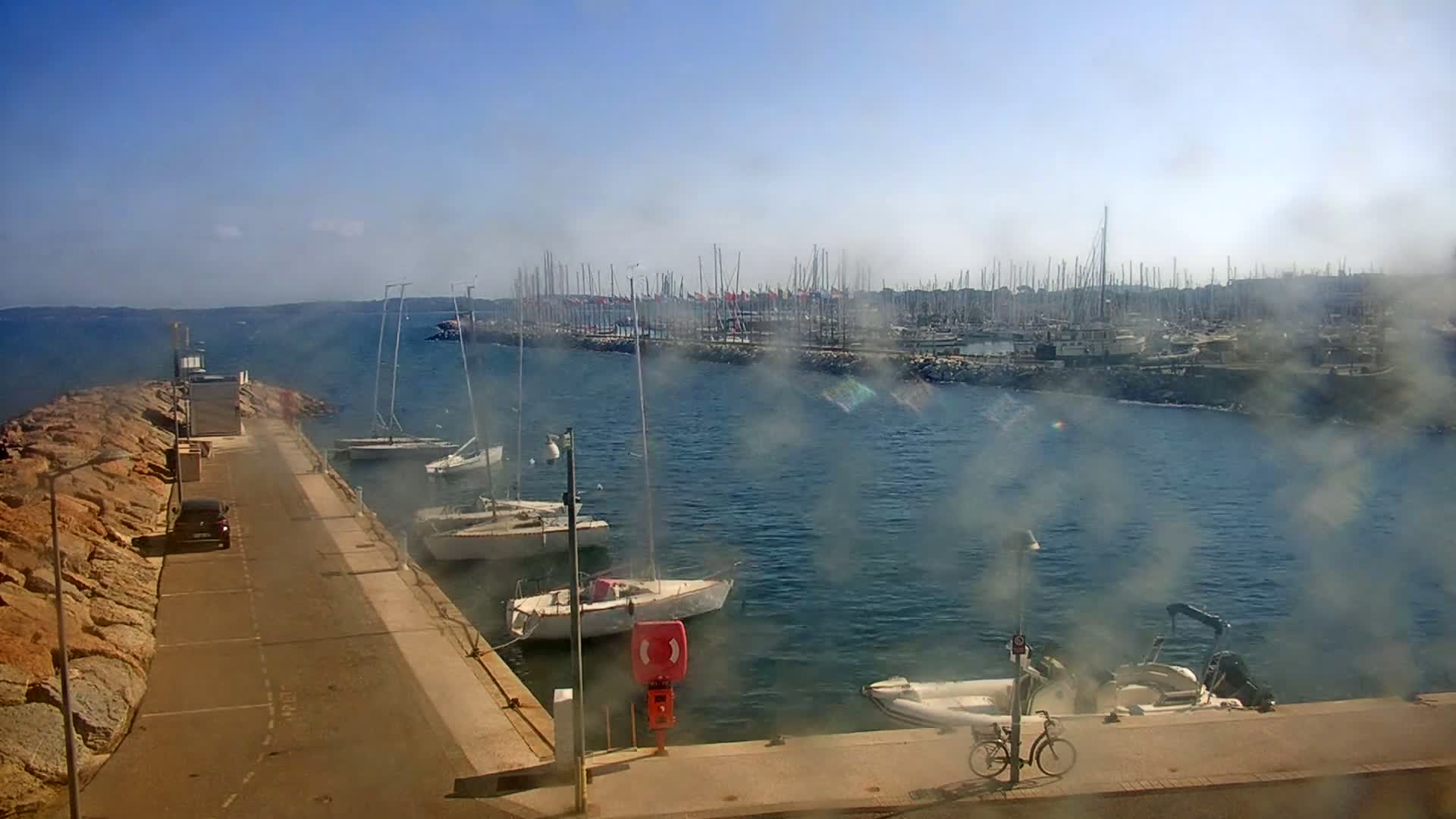 A hazy, sunny day reveals a harbor with several sailboats moored along a rocky shore, a few additional boats near a paved walkway, and a car parked nearby.