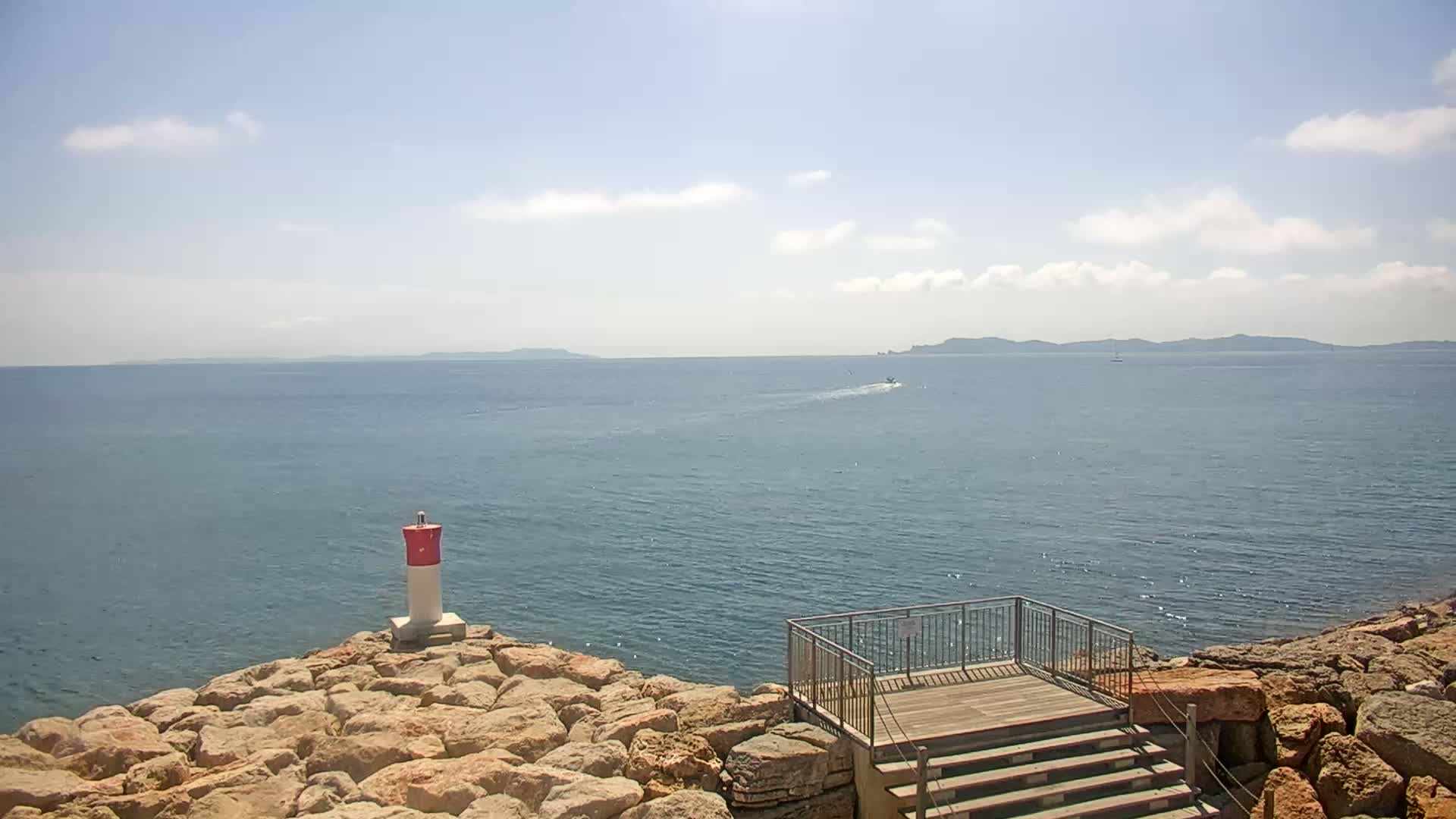 A rocky shoreline with a small lighthouse and viewing platform overlooks a calm sea under a partly cloudy sky, with distant islands visible on the horizon.