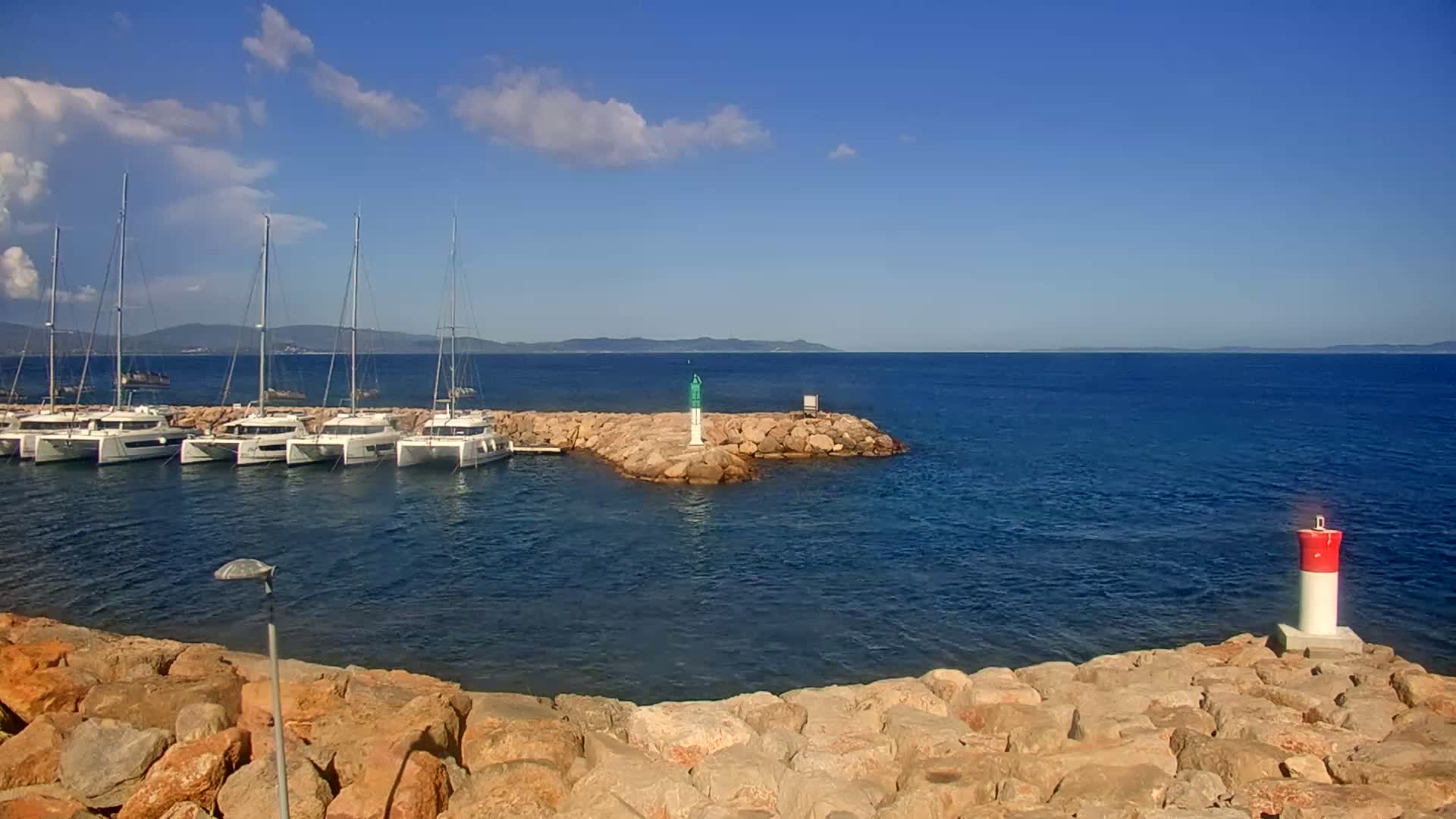 Several sailboats are moored in a calm harbor, with rocky breakwaters and buoys visible under a mostly clear blue sky.