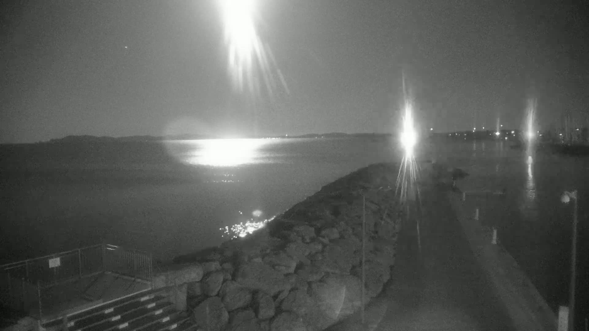 A nighttime, low-light view of a rocky shoreline and calm body of water reflecting bright lights across the surface, under a clear sky.