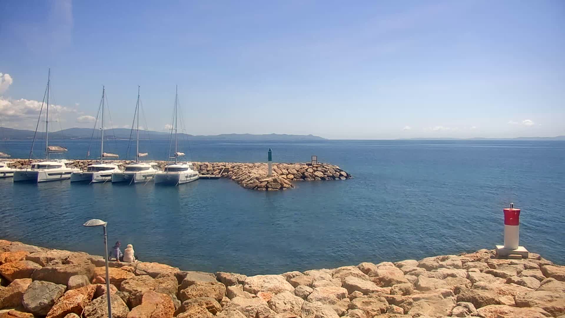 Several sailboats are moored in a calm, clear blue ocean next to a rocky shore where two people sit on the rocks under a bright sunny sky.