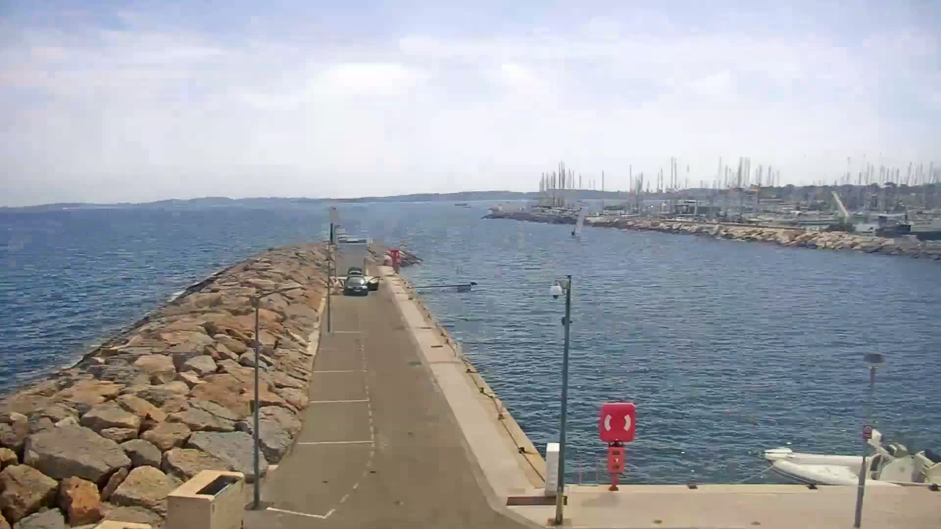 A paved road bordered by a rock wall leads to a harbor filled with sailboats under a partly sunny sky.