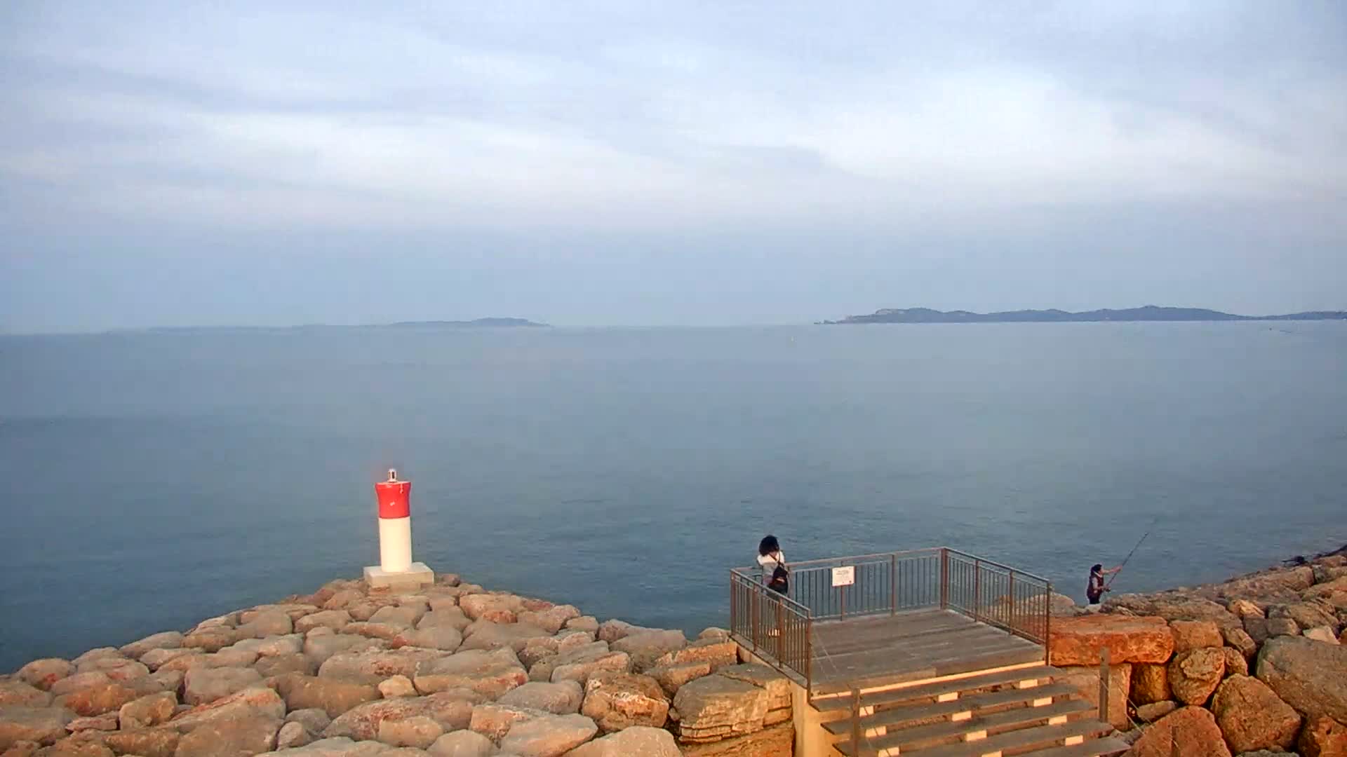 A rocky shoreline overlooks a calm sea under a pale sky, with a small lighthouse, a viewing platform, and two people—one standing and one fishing.