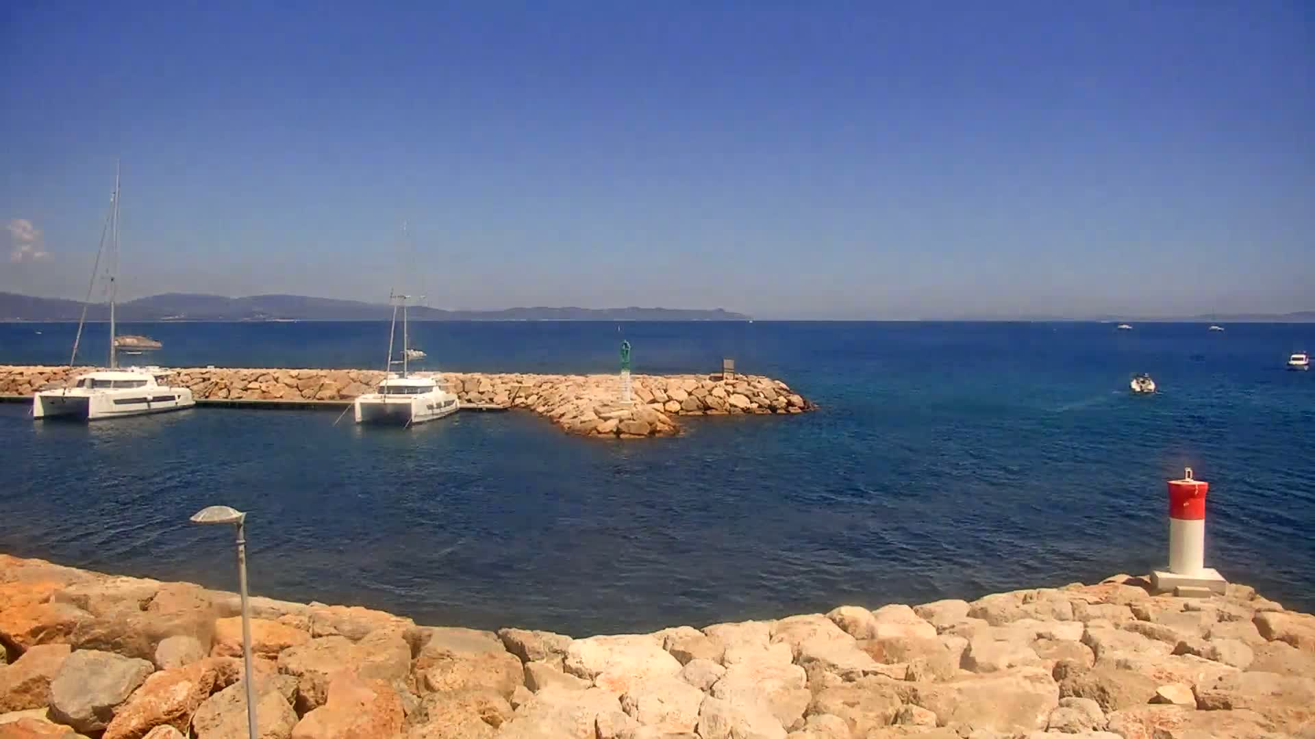 Two sailboats are moored in a calm, clear blue sea near a rocky breakwater on a sunny day, with distant mountains visible on the horizon.