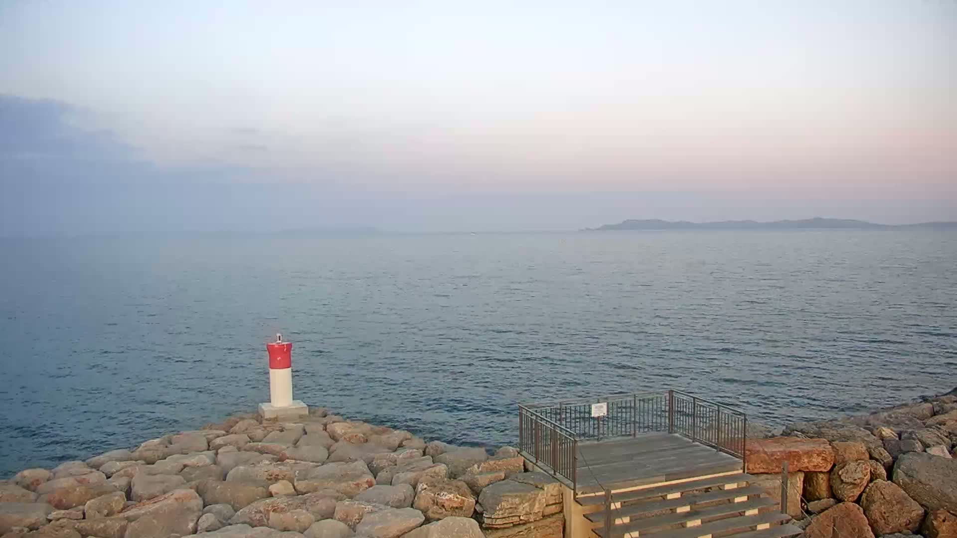 A red and white marker sits on a rocky shoreline overlooking a calm sea under a hazy, pale sky.