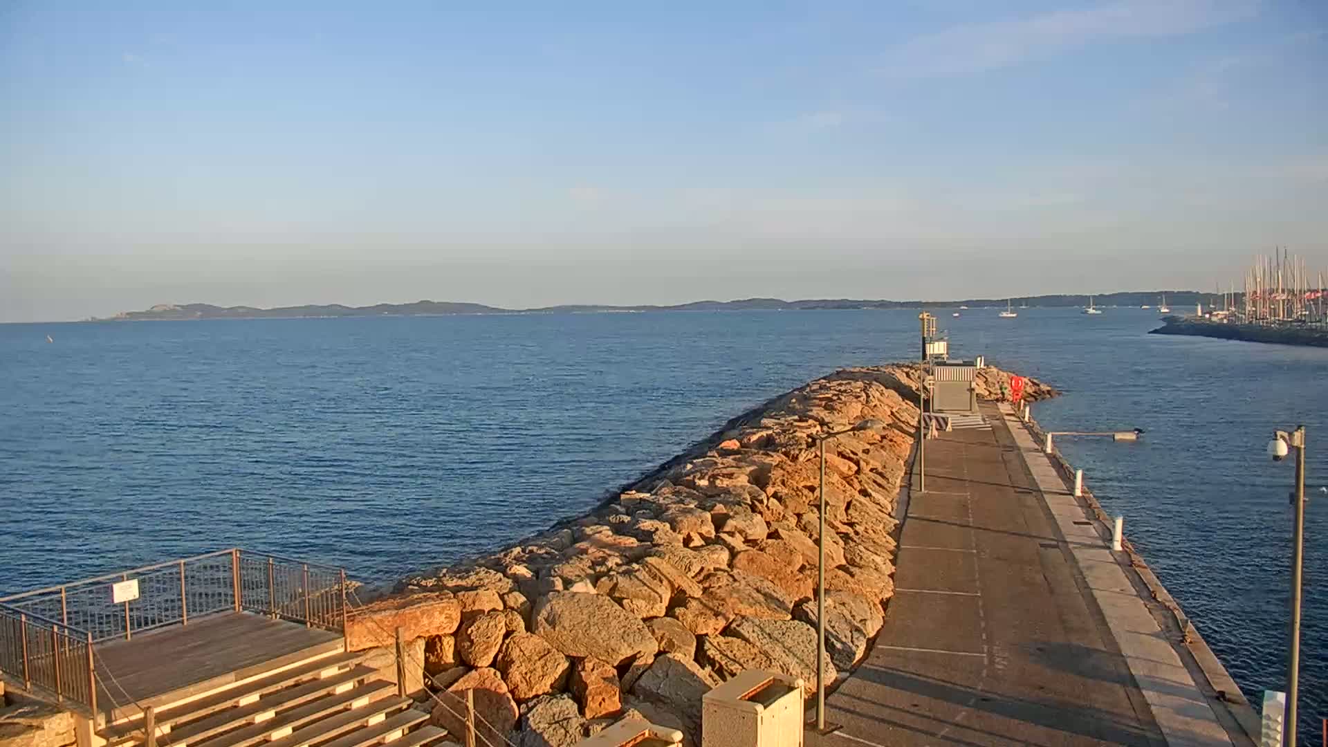 A rocky breakwater extends into a calm, blue sea under a clear sky, with a distant shoreline and several sailboats visible in the background.