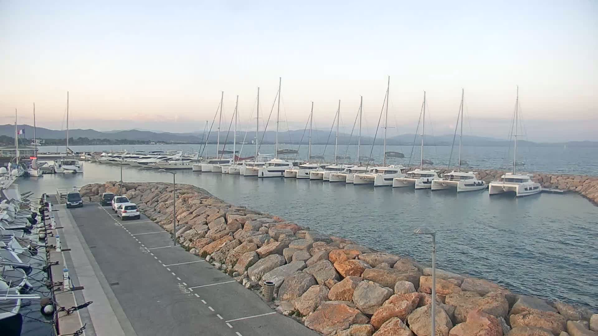A calm harbor filled with numerous sailboats and catamarans, bordered by a rocky wall and a paved area with cars parked nearby, under a pale sky.