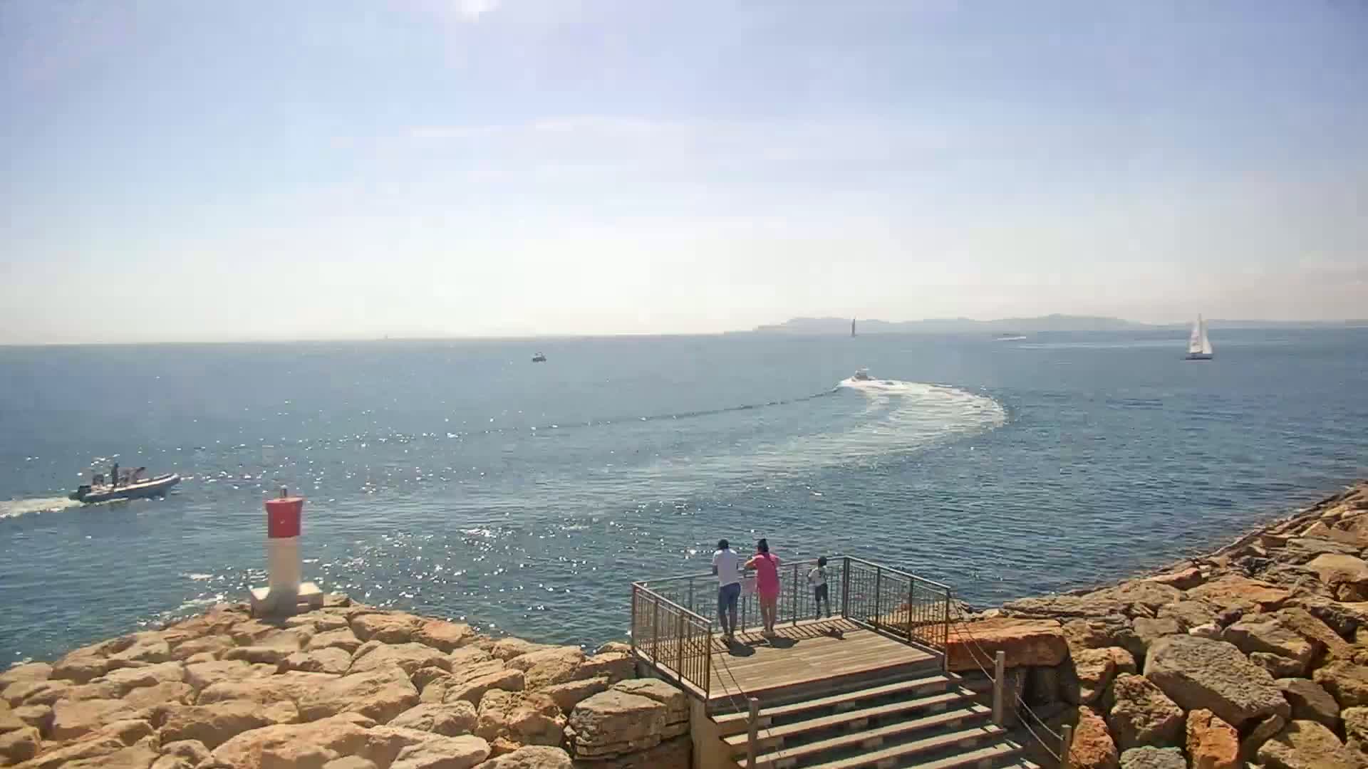 A sunny day at sea shows a family standing on a wooden viewing platform overlooking a rocky coastline with several boats and a sailboat in the distance.
