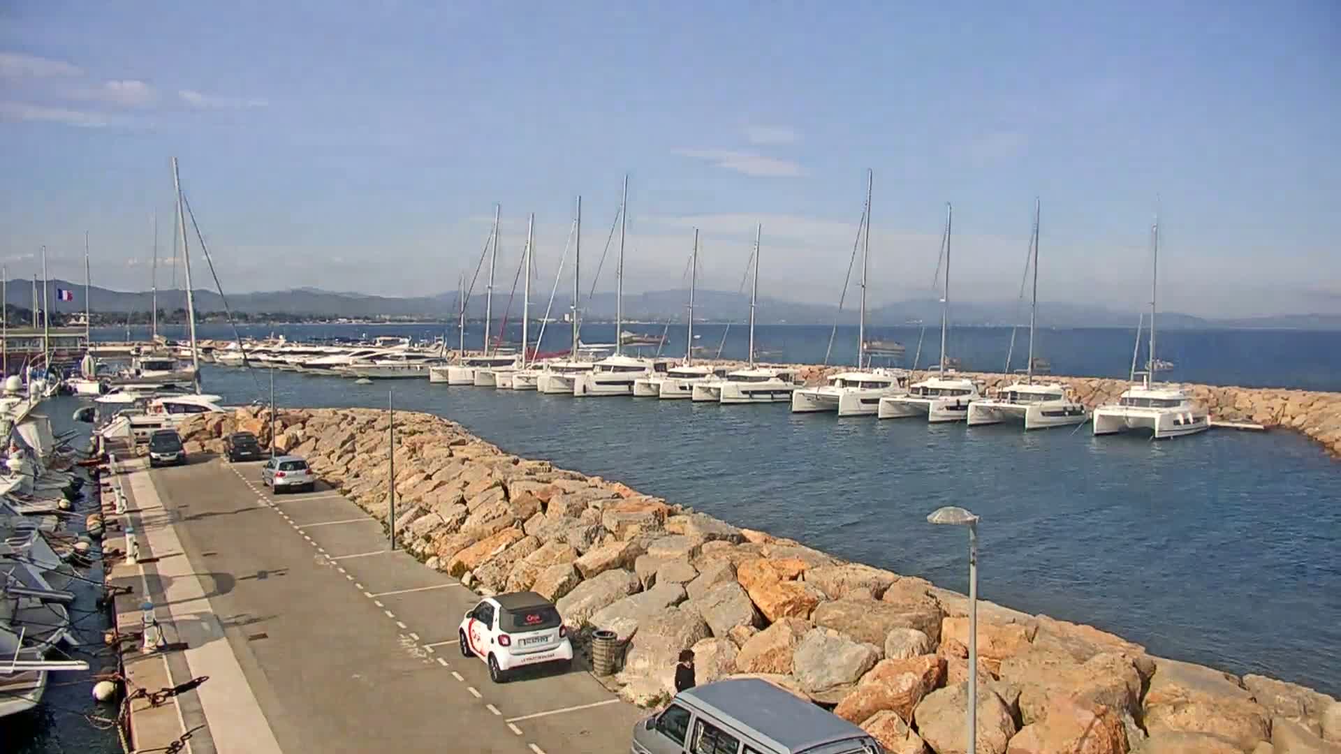 A marina with numerous sailboats and catamarans is situated beside a rocky shore on a sunny day.