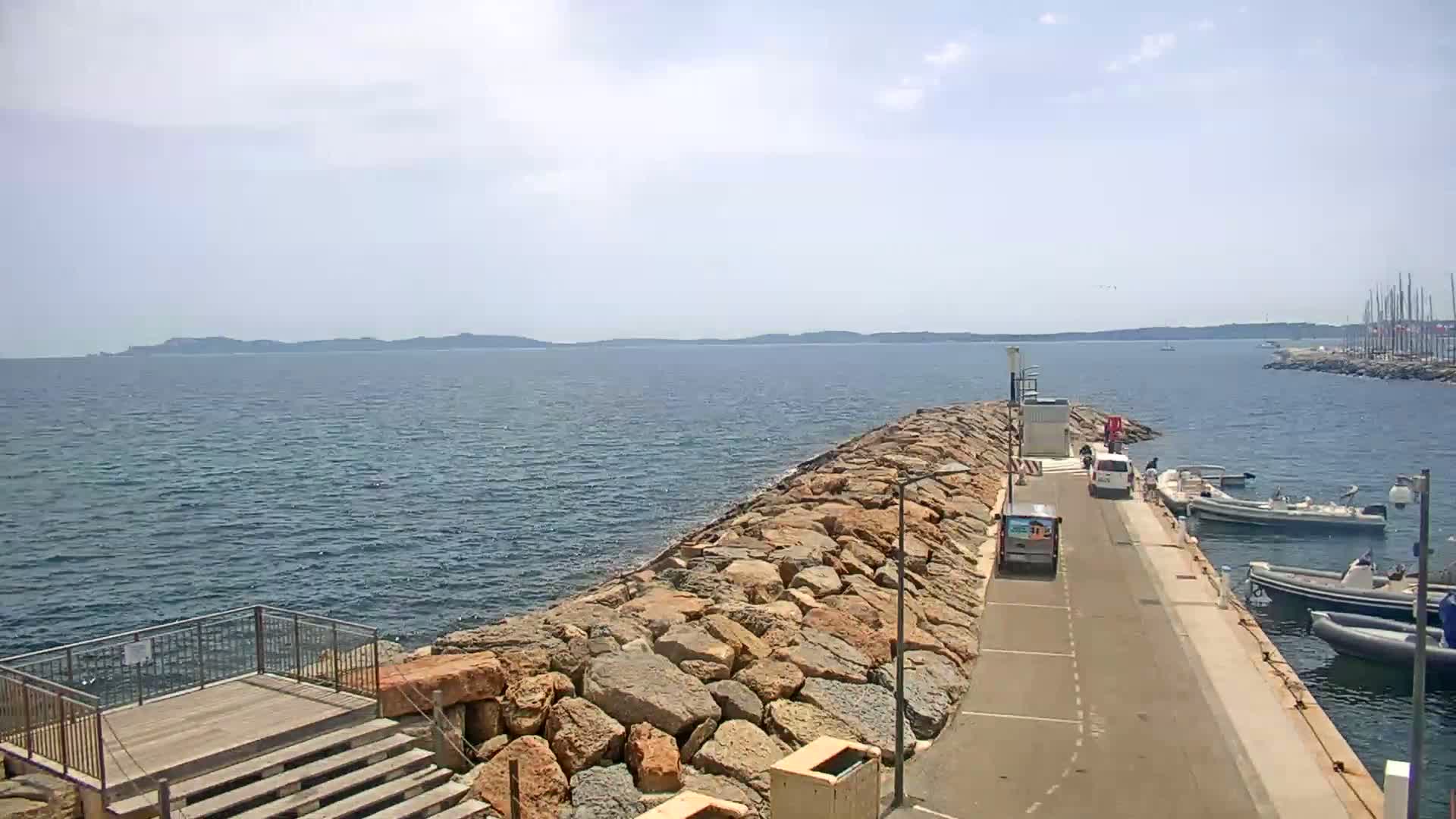 A paved road extends from a rocky breakwater into a calm sea under a partly cloudy sky, with several small boats moored nearby and a distant shoreline visible.
