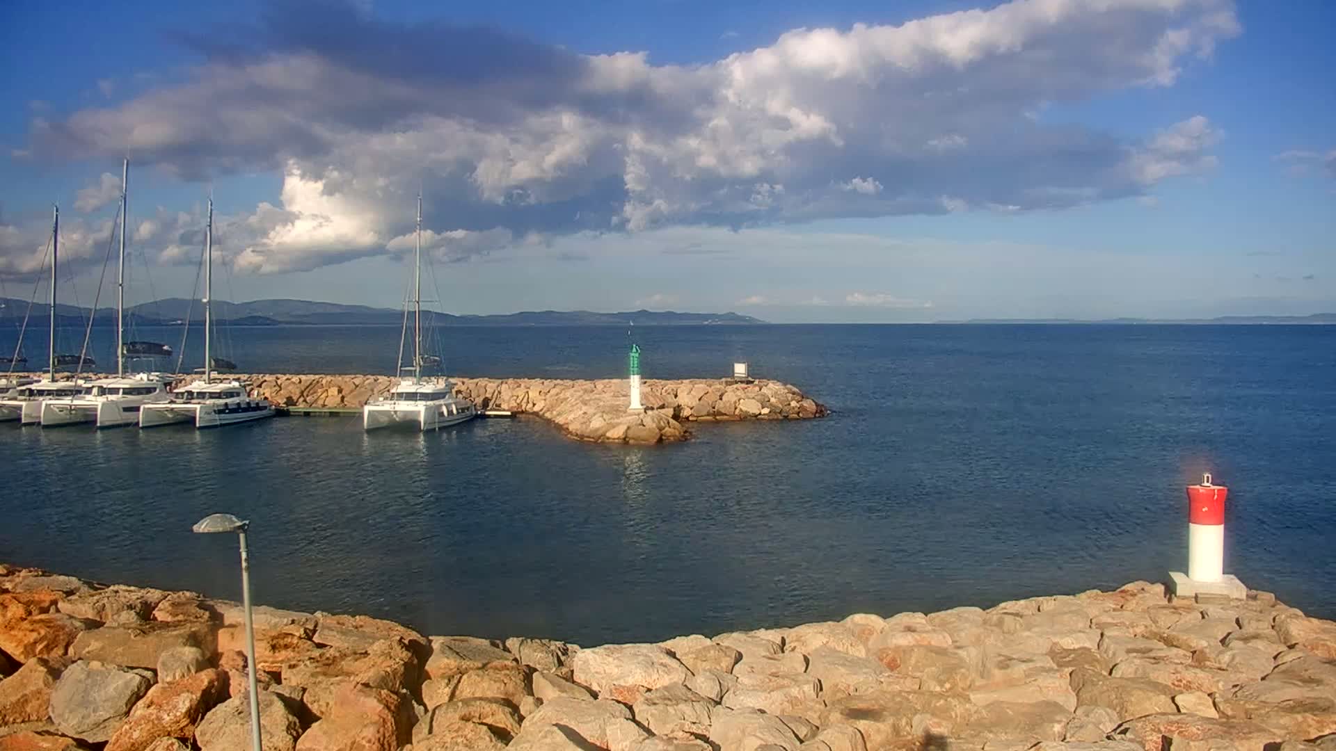 Several sailboats are moored in a calm harbor, partially sheltered by a rocky breakwater under a partly cloudy sky.