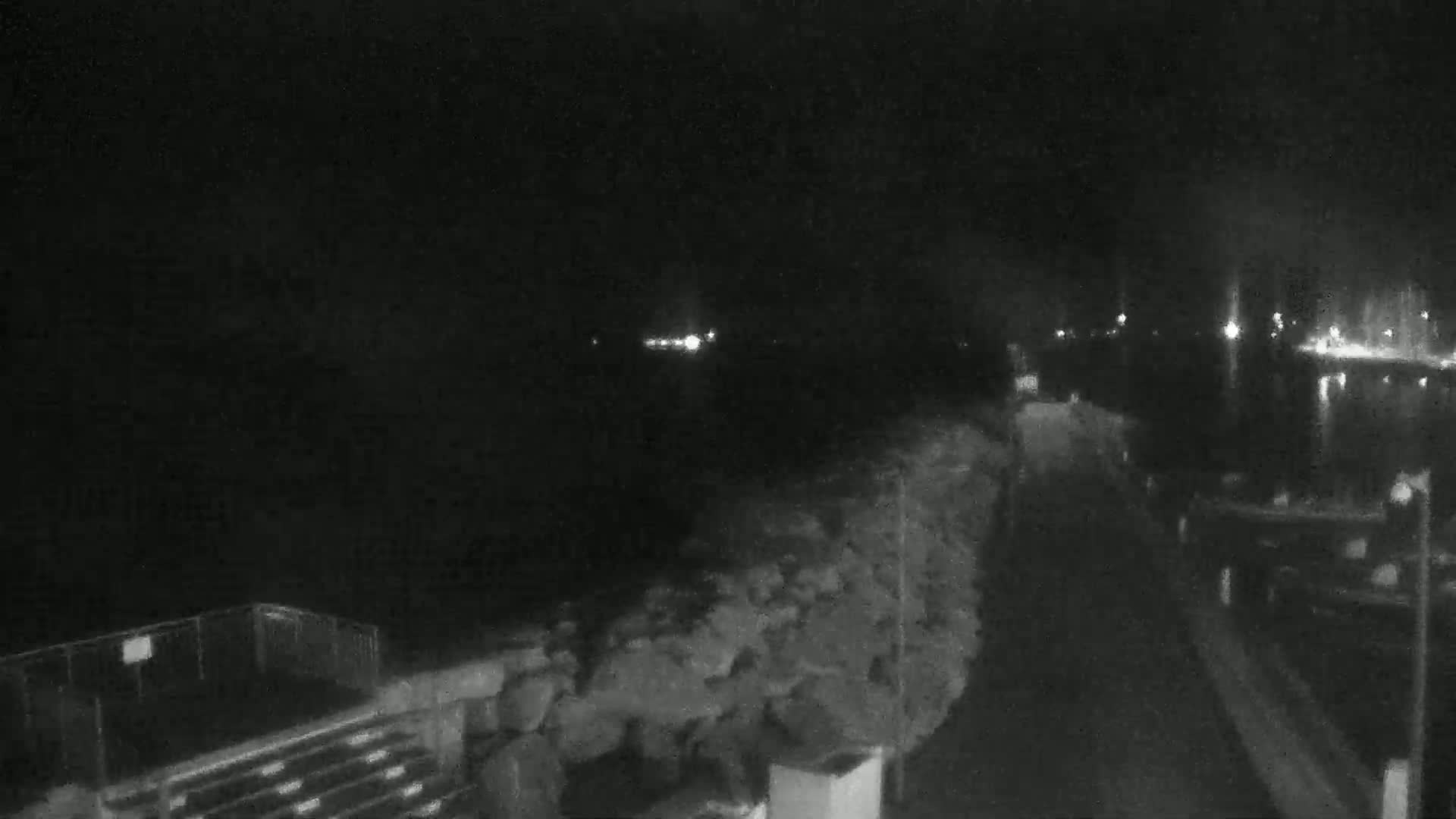 A nighttime, low-light view of a dark waterfront area with a rocky embankment, a paved walkway, and distant lights across the water.