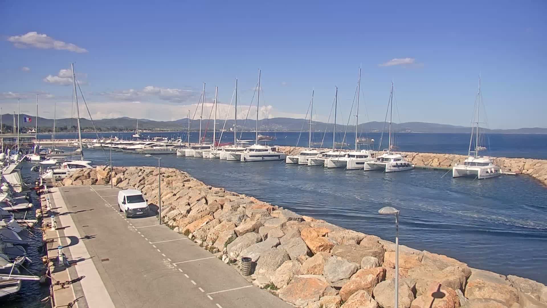 A marina filled with sailboats and catamarans is nestled next to a rocky shore under a mostly sunny sky.
