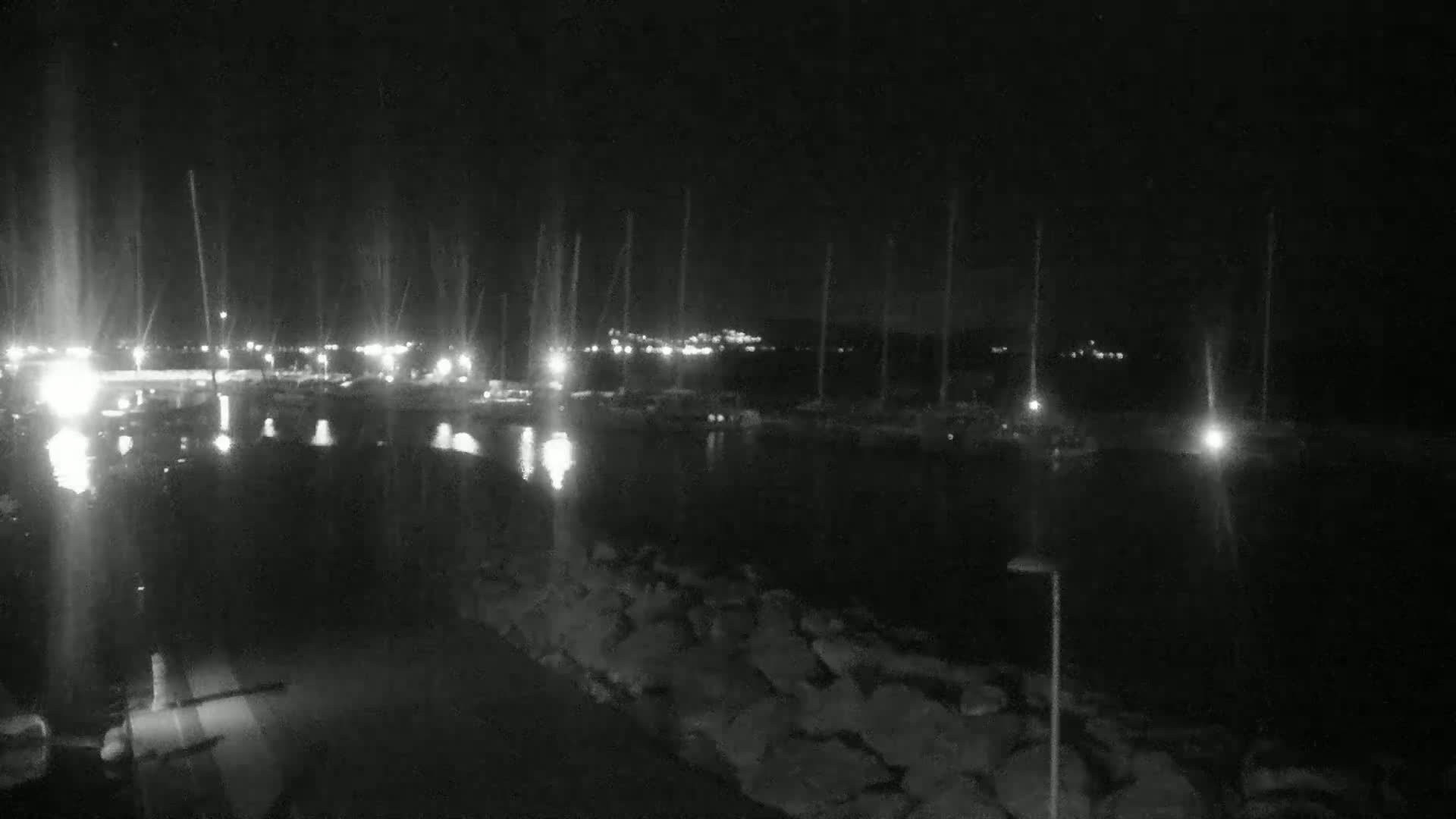 A nighttime view of a harbor filled with boats, illuminated by scattered lights, with a rocky shoreline in the foreground under a dark sky.