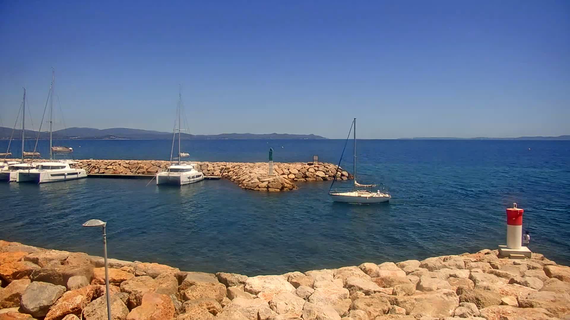 Several sailboats are moored in a calm, blue sea near a rocky shore under a clear, sunny sky.