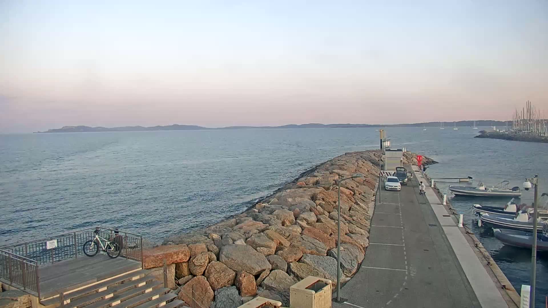 A rocky breakwater extends into a calm sea under a pale, dusky sky, with several boats moored alongside a paved walkway and a bicycle leaning against a railing.