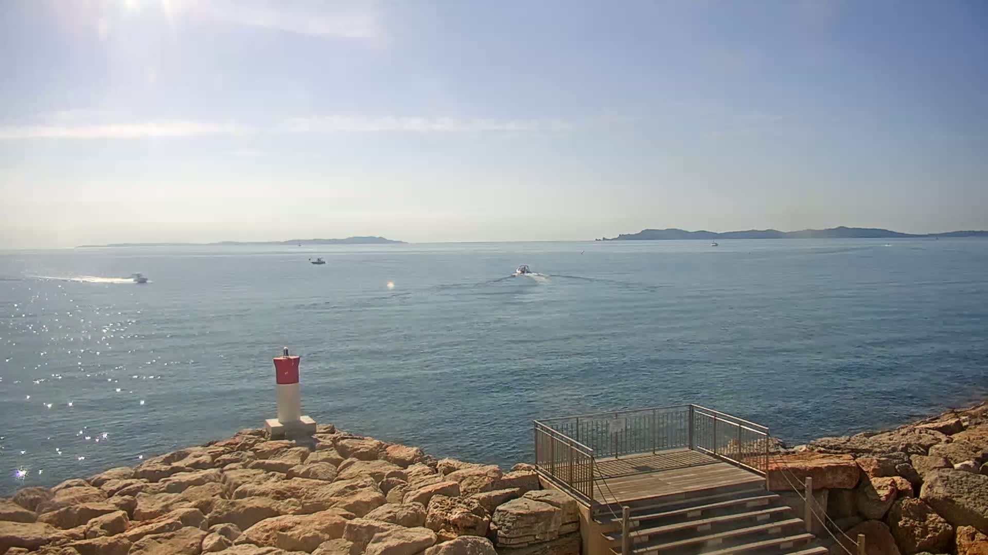 A rocky shoreline with a small viewing platform overlooks a calm, sunny sea with several boats and distant islands.