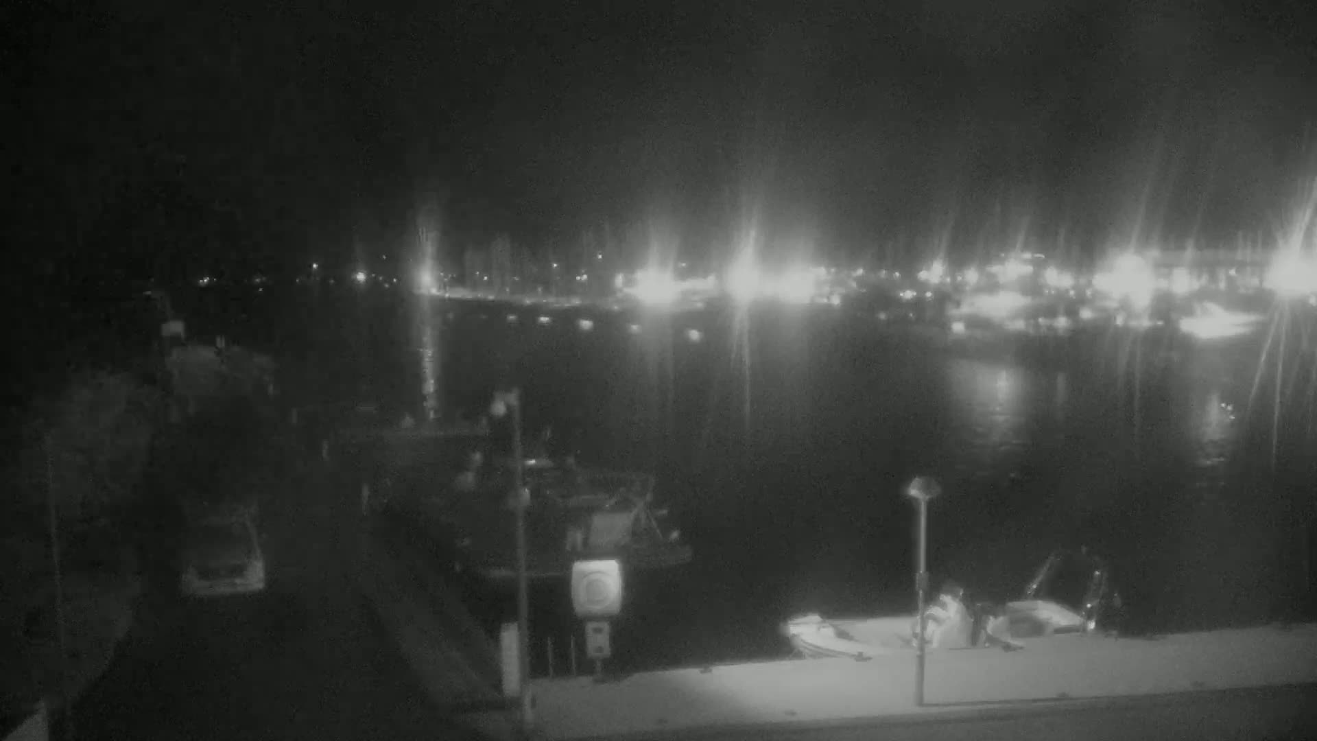 A nighttime view of a harbor with boats docked, cars parked nearby, and lights illuminating the water and distant shoreline.