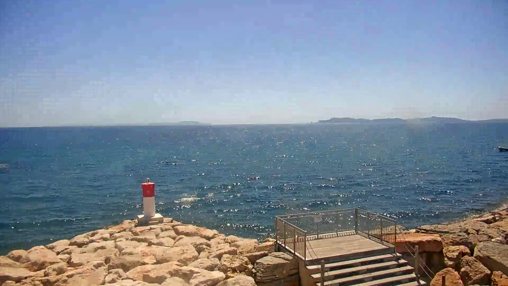 A rocky shoreline with a small viewing platform overlooks a calm, sparkling blue sea under a clear, sunny sky, and a distant landmass is visible on the horizon.