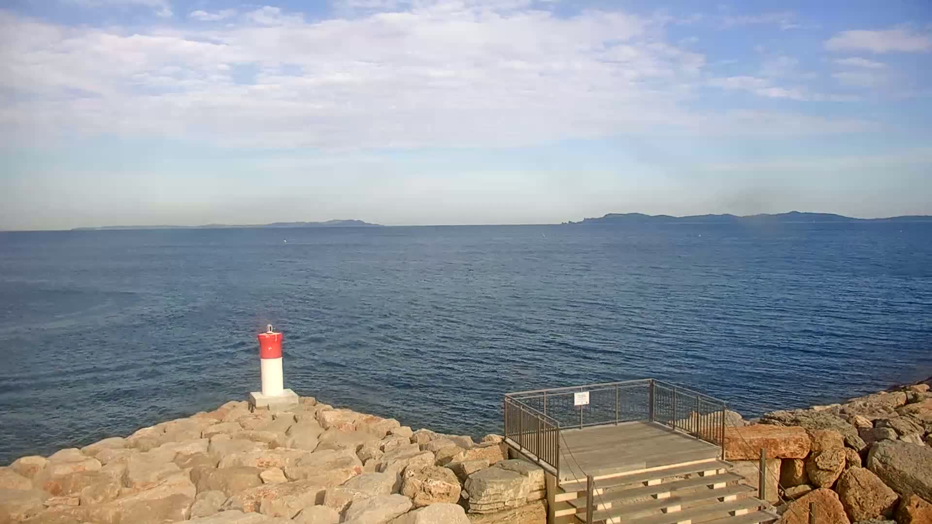 A red and white buoy stands on a rocky outcrop overlooking a calm sea under a partly cloudy sky, with distant islands visible on the horizon.