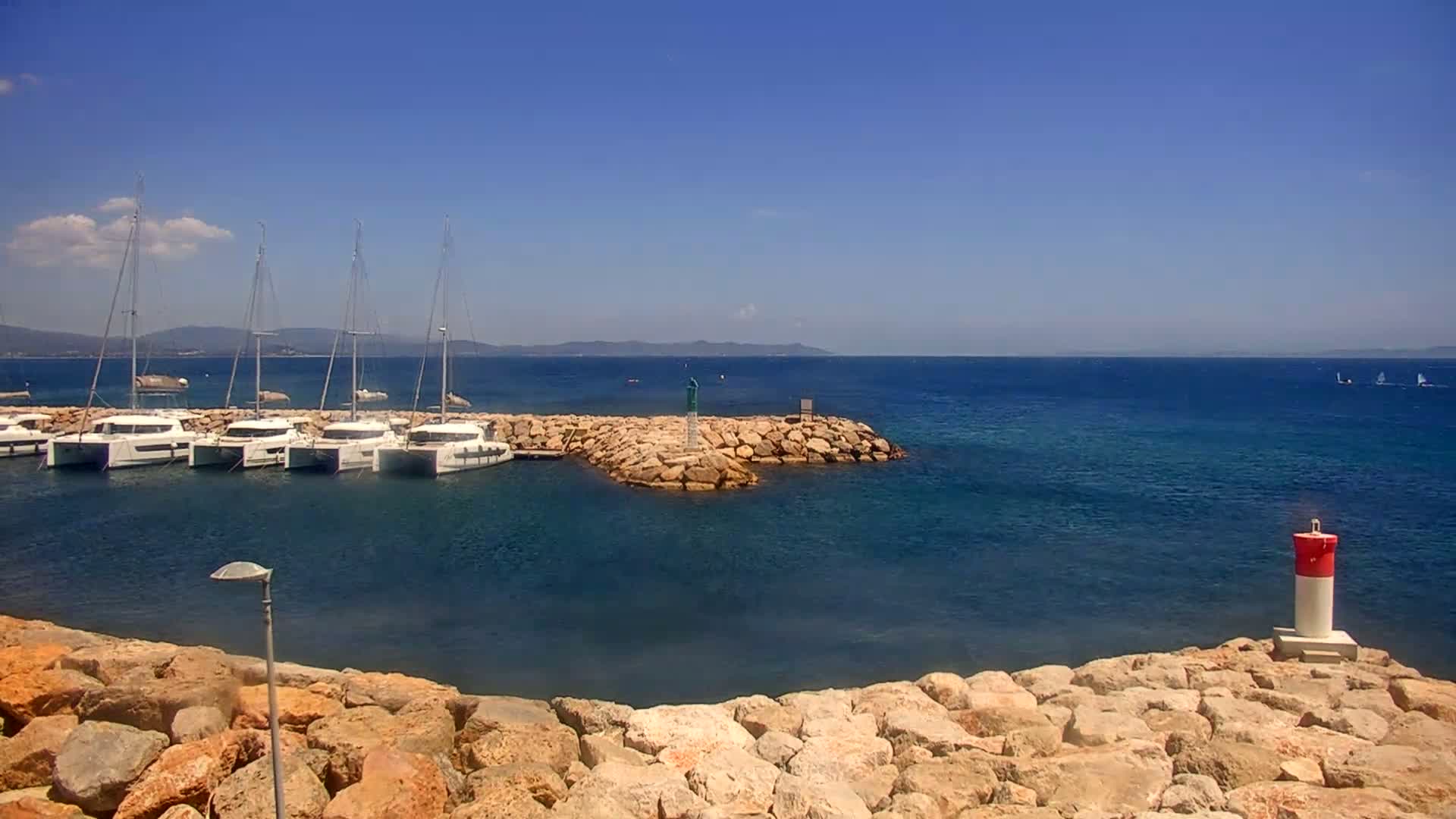 Several sailboats are moored in a calm harbor protected by a rock jetty under a clear blue sky.