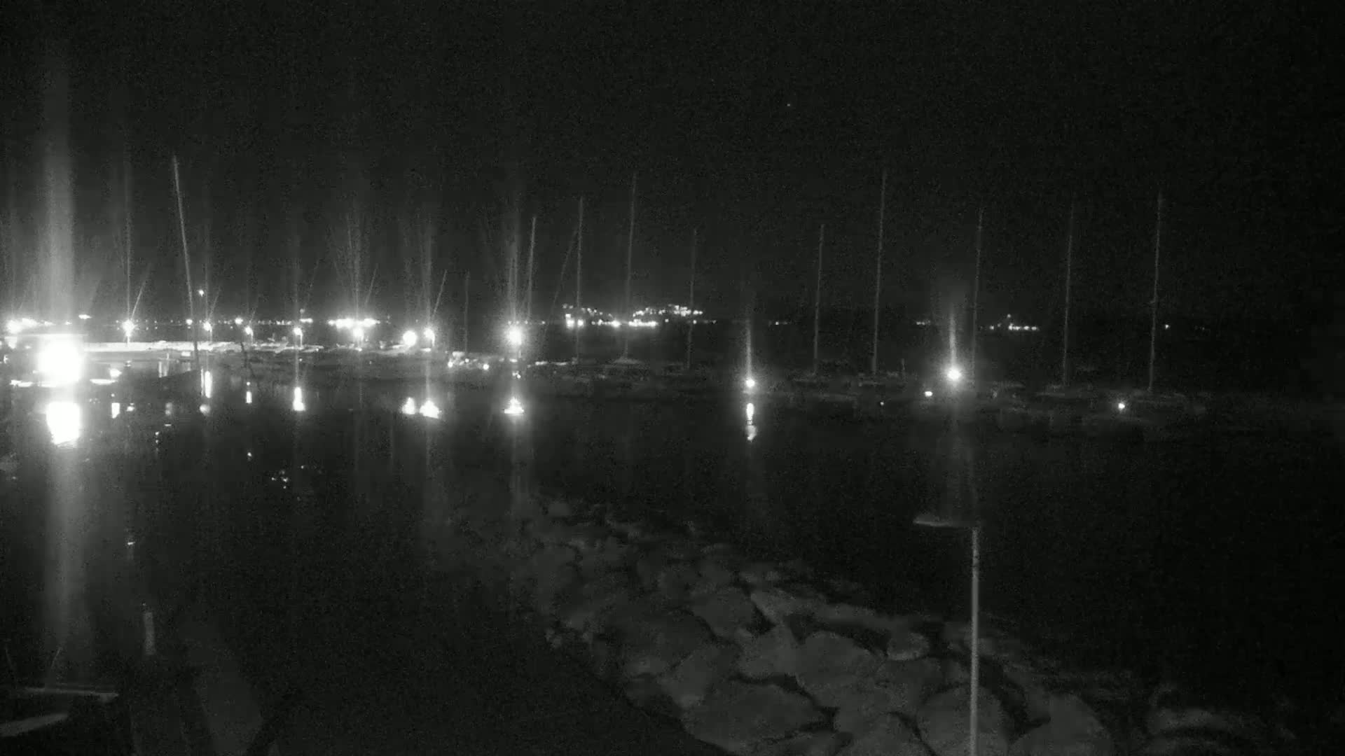 A nighttime view of a harbor filled with boats, illuminated by lights from the docks and vessels, with a rocky shoreline in the foreground.