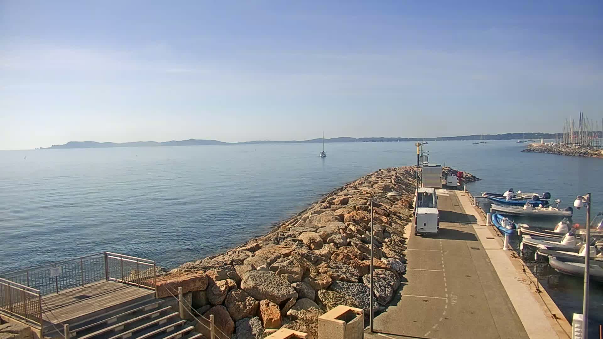 A rocky breakwater extends into a calm, blue sea under a clear, sunny sky, with several boats moored alongside a paved walkway.