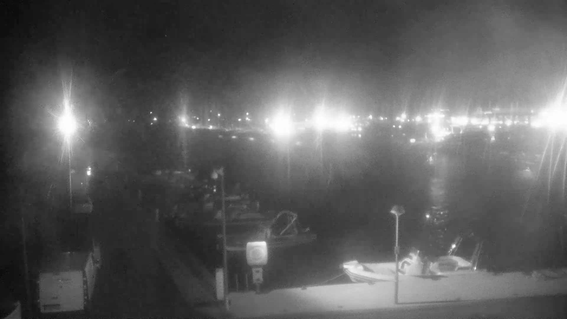 At night, several boats are docked at a marina, with city lights visible in the distance under a dark sky.