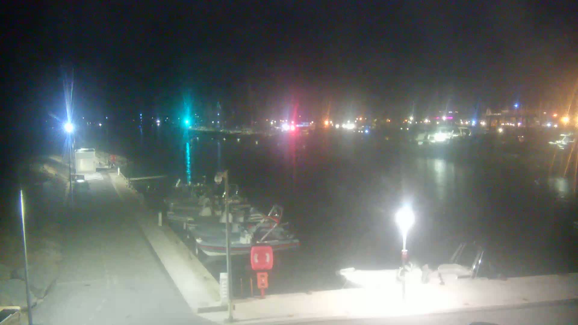 At night, a calm body of water is filled with boats, and a dock extends into the water with a few more boats moored alongside.