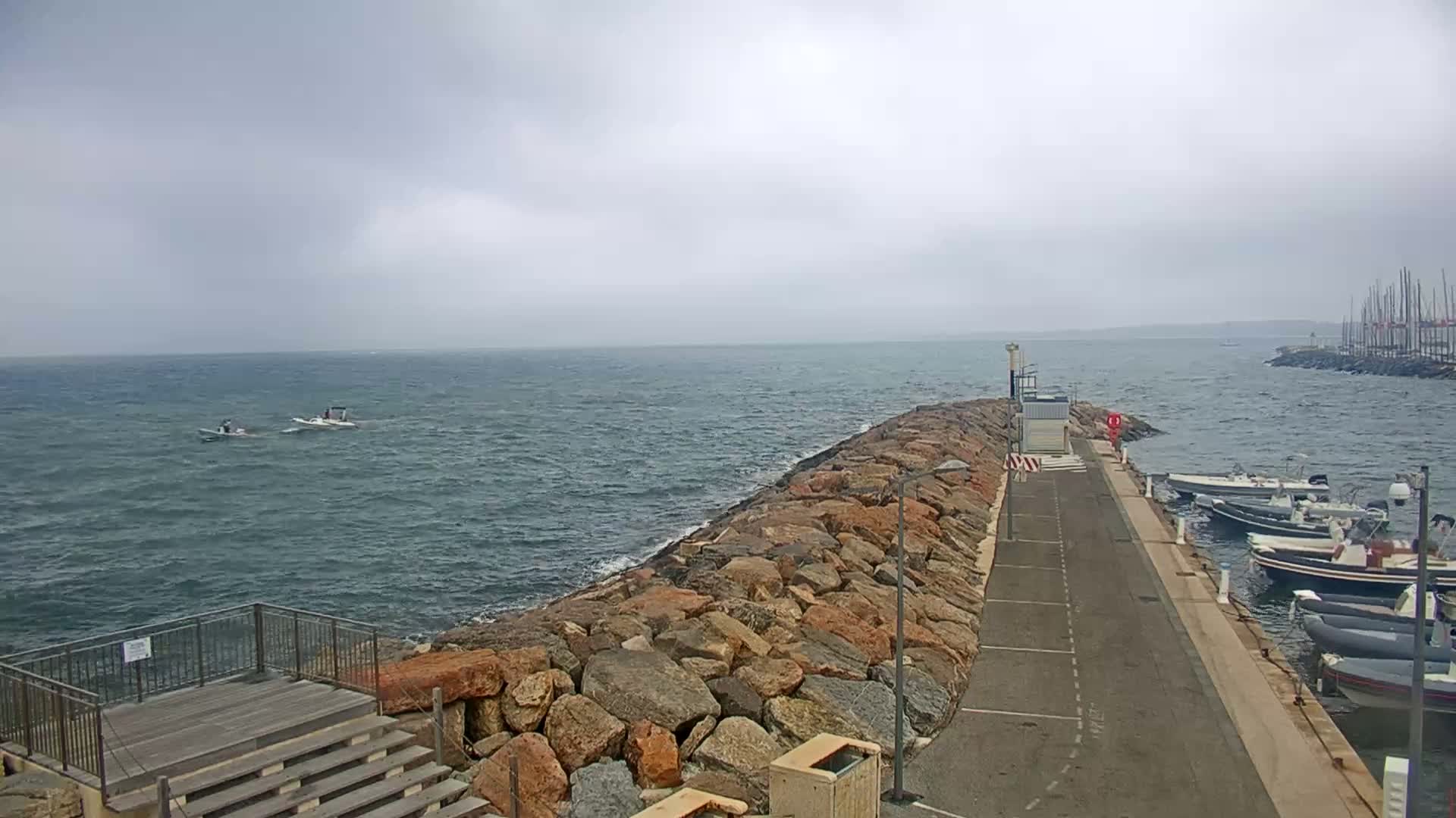 A rocky breakwater extends into a choppy sea under a cloudy sky, with several small boats visible on the water and a marina with moored motorboats in the background.